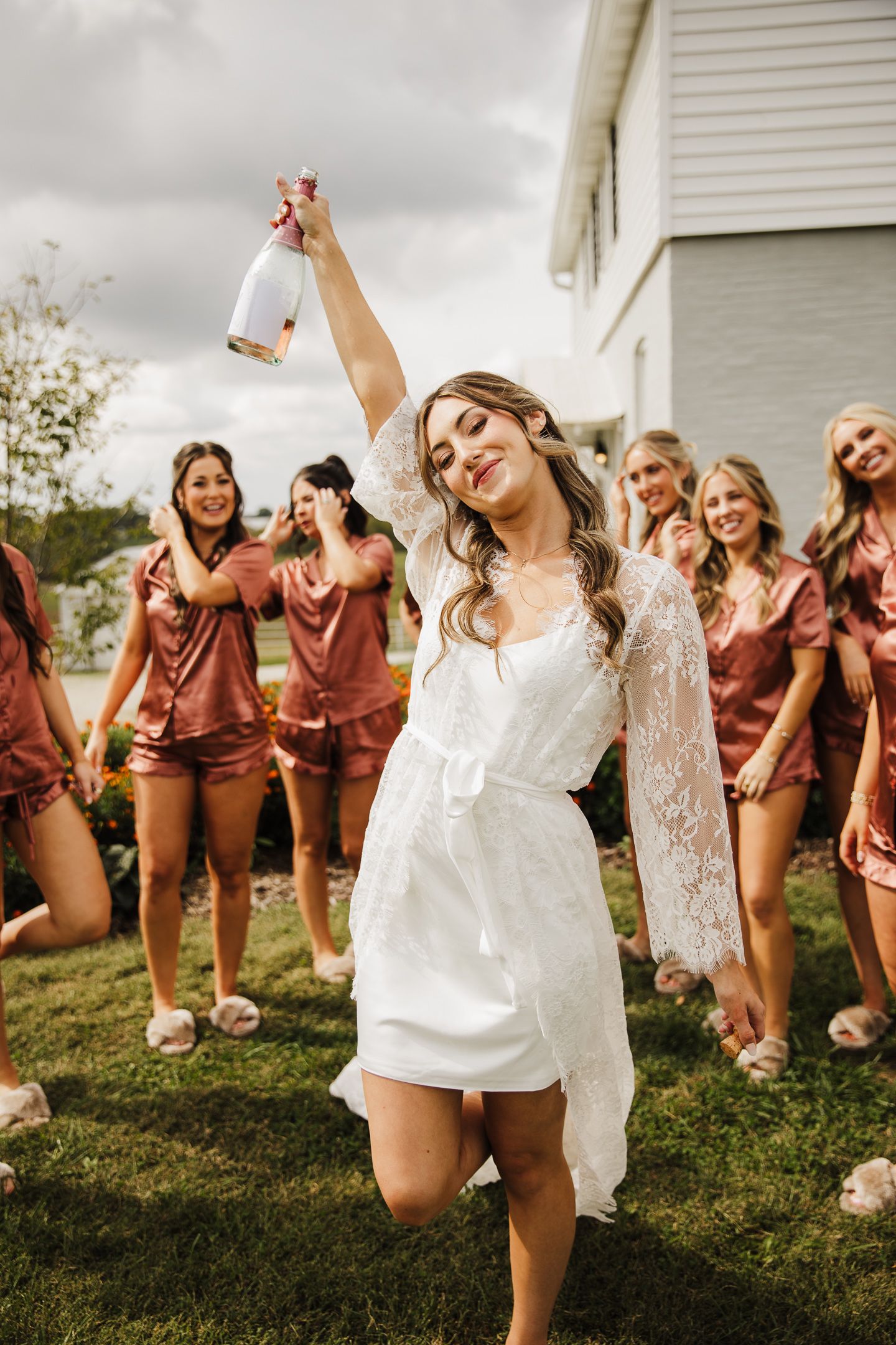 Bridesmaids in burgundy dresses pose playfully on grass during a wedding photo session.