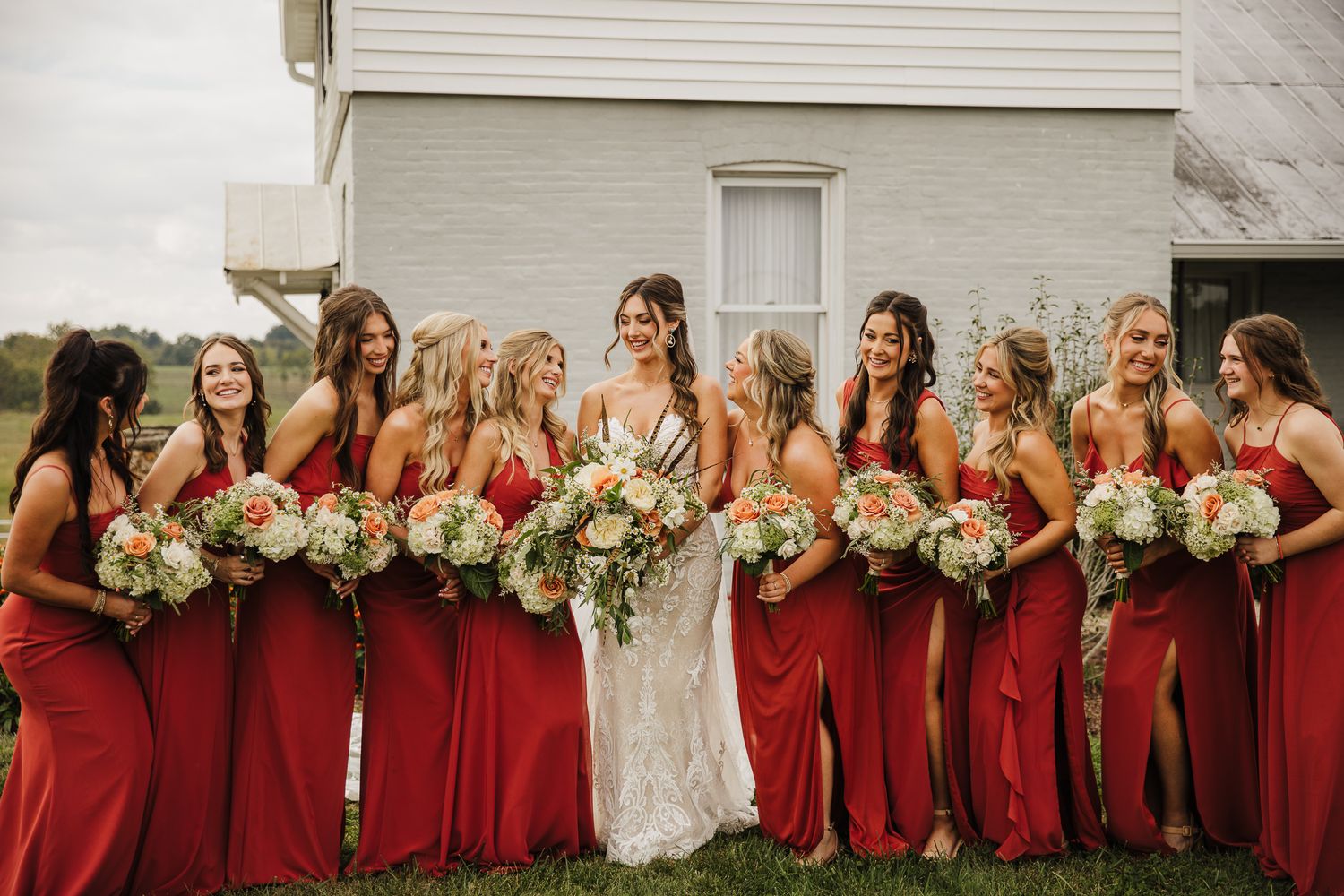 A wedding party in red bridesmaid dresses poses outdoors with floral bouquets beside a white building.