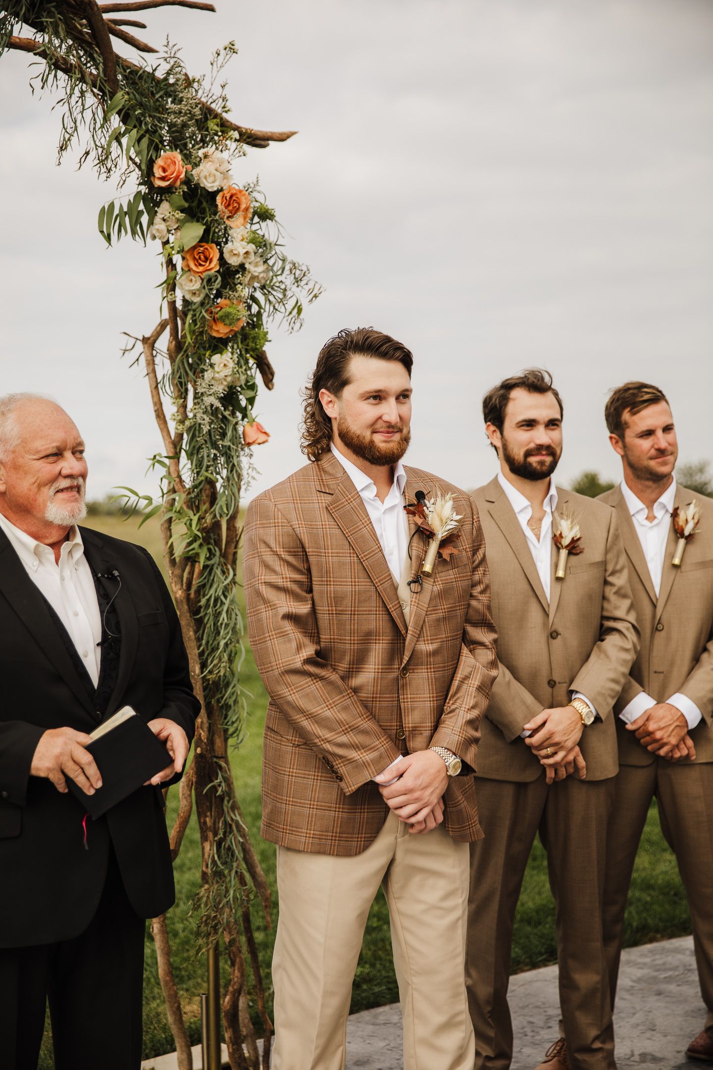 Wedding party in earth-toned suits poses outdoors under floral arch.