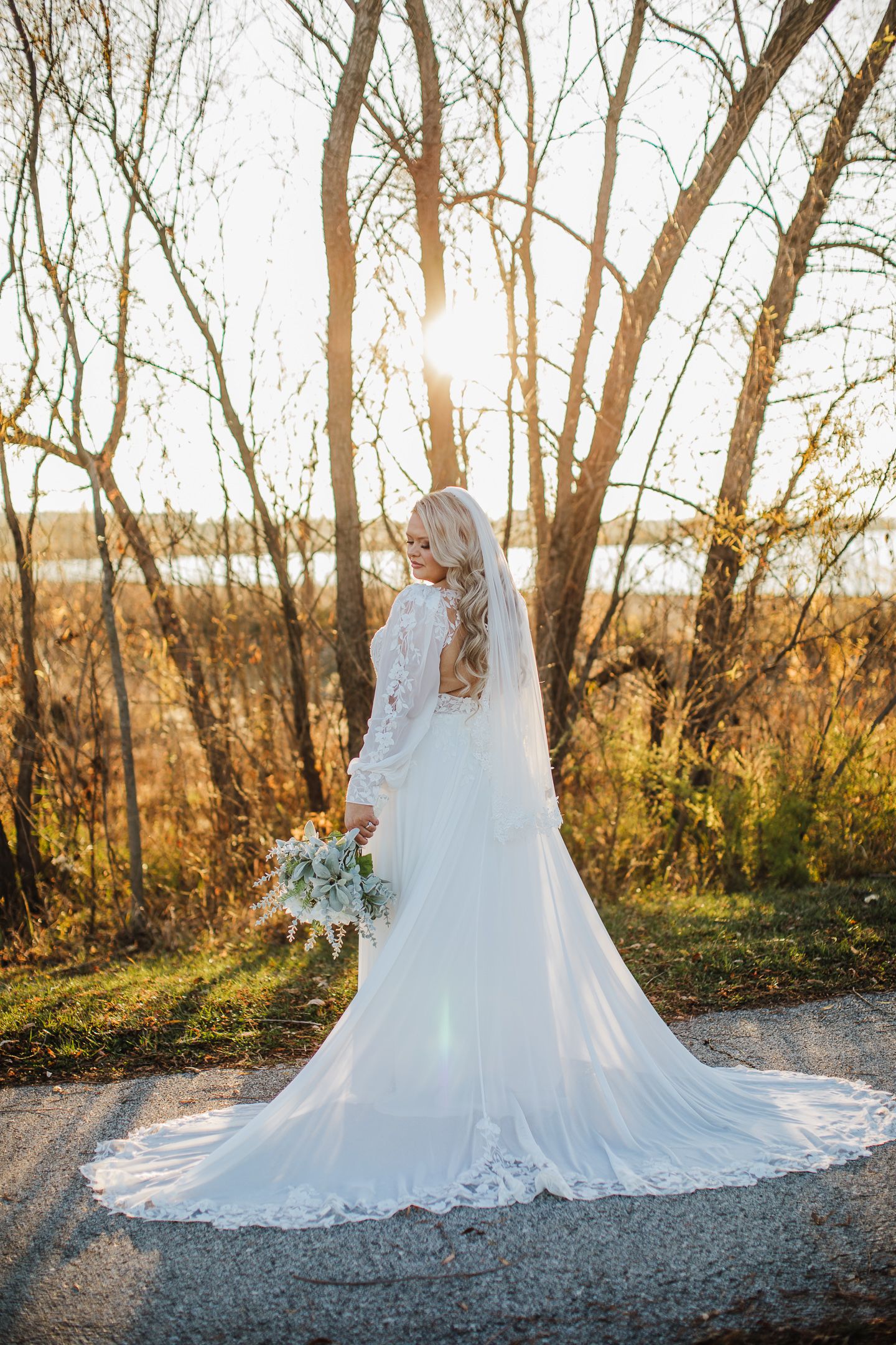 A bride in a flowing white lace gown poses against sunlit trees.