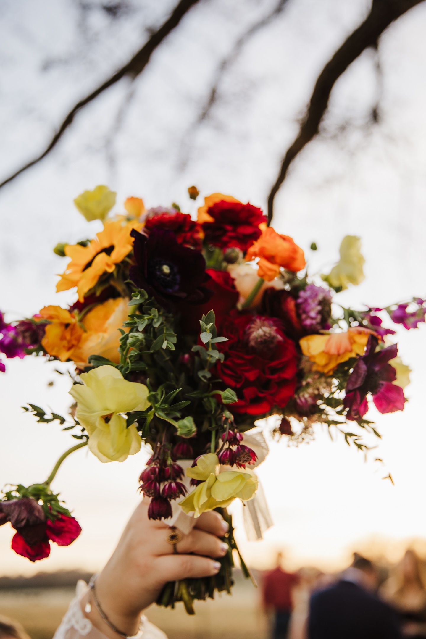 A colorful floral arrangement featuring red, yellow, and orange flowers is held up against a bright sky background.