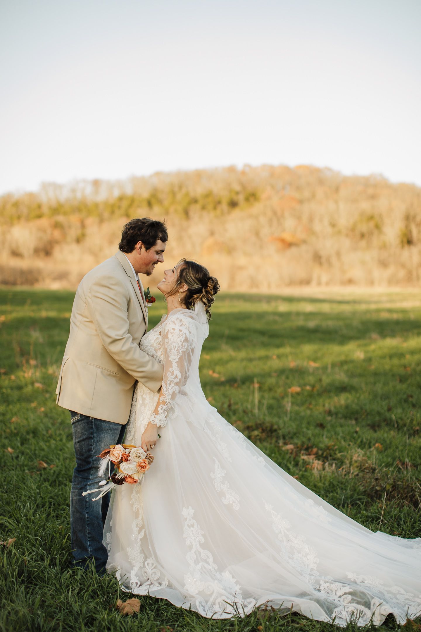 Wedding couple sharing intimate moment in outdoor field with bride's lace dress trailing behind them.