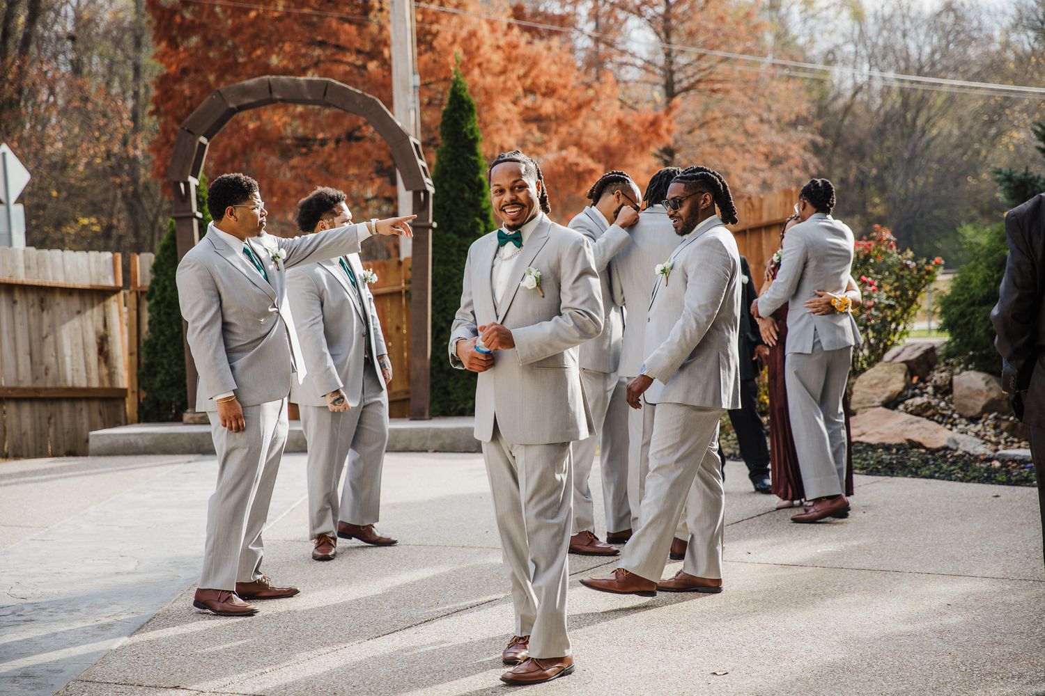 Groomsmen in light gray suits gather outdoors during autumn wedding celebration.