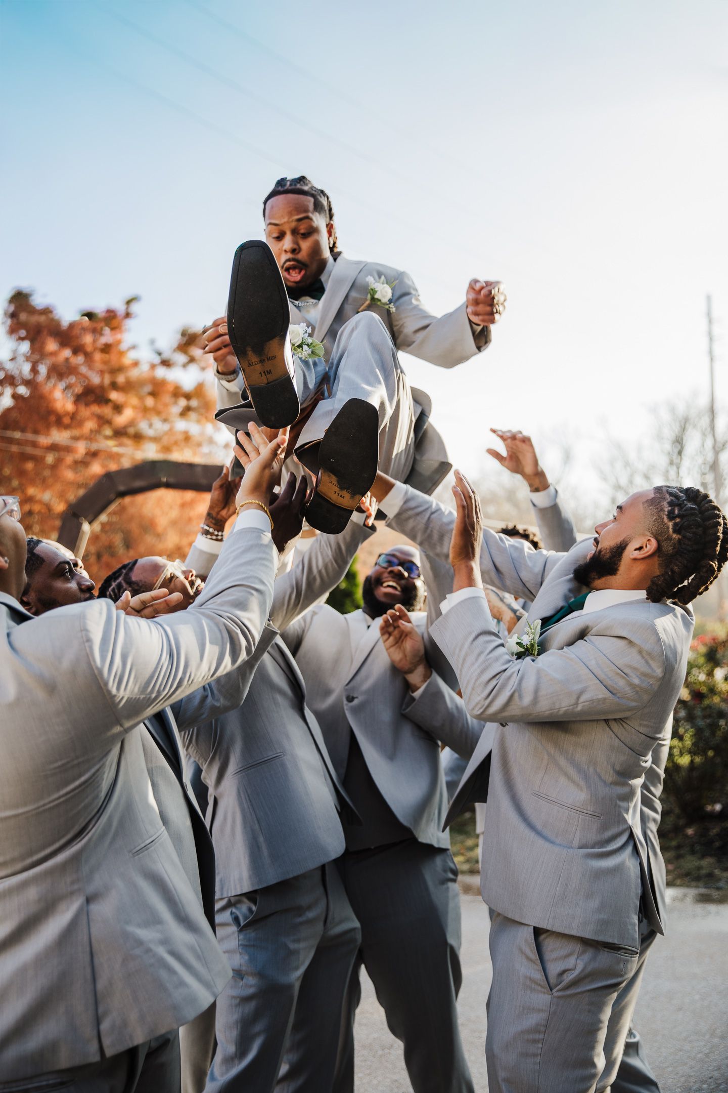 Groomsmen in gray suits lift and celebrate during outdoor wedding festivities.
