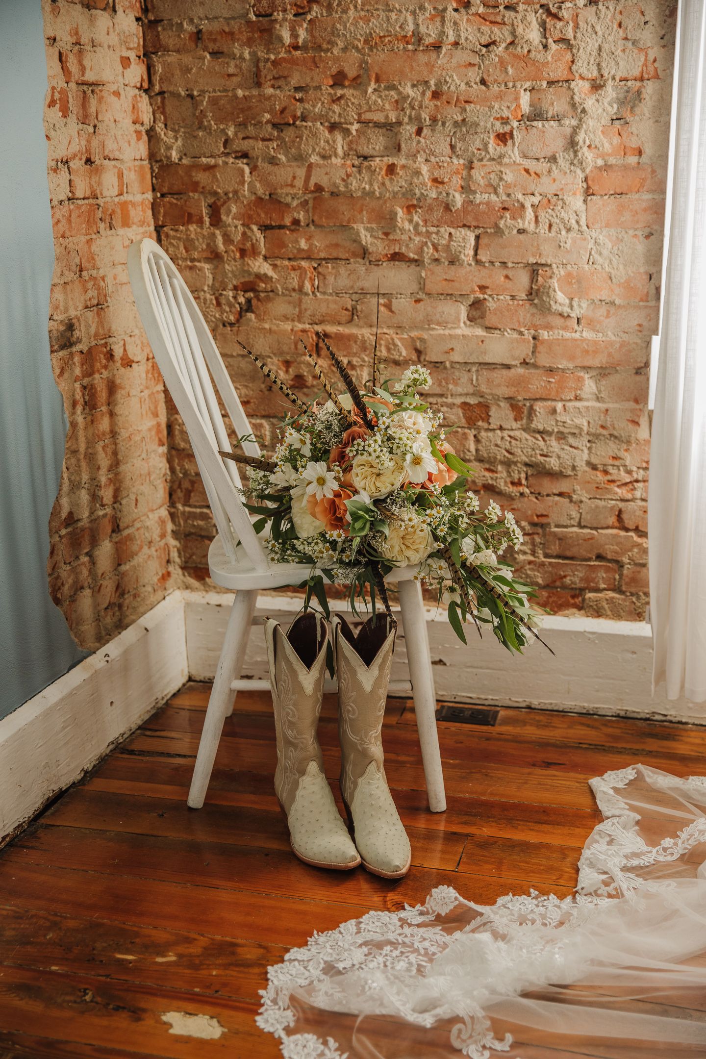 A rustic wedding setup with cowboy boots, floral bouquet and white chair against exposed brick wall.