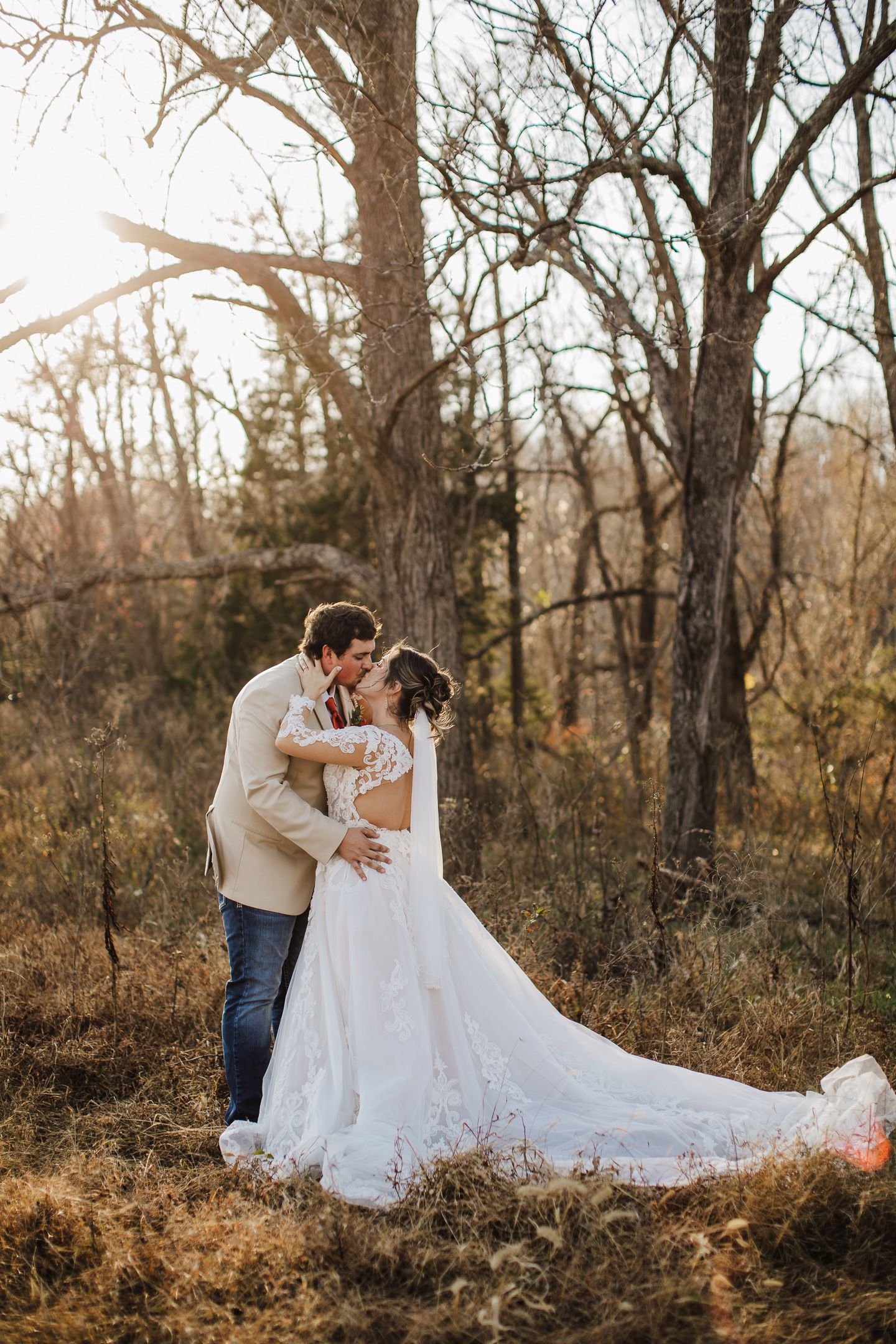 A romantic wedding couple embraces in a sunlit autumn forest setting with golden light filtering through bare trees.