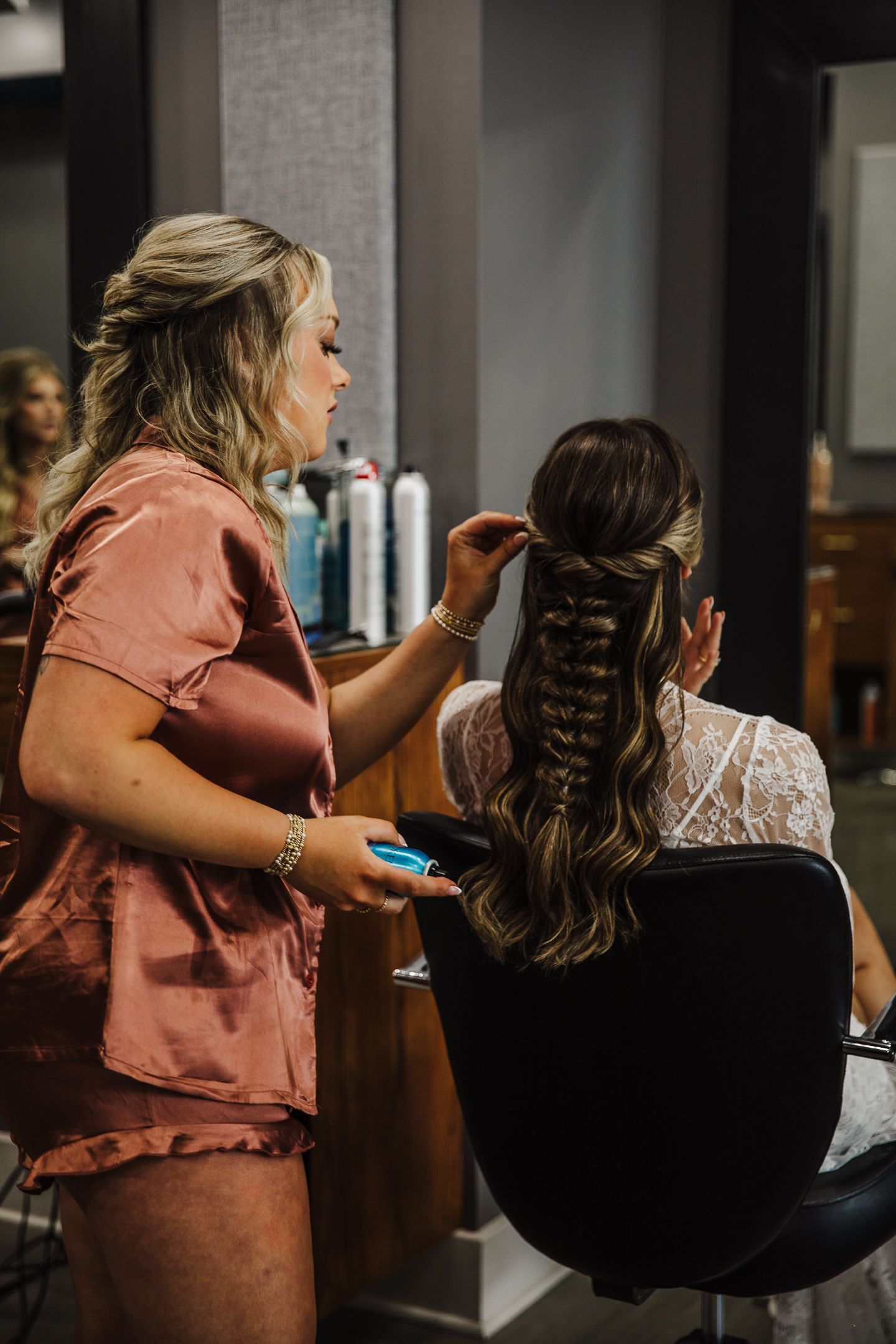 A stylist in coral scrubs arranges an intricate braided hairstyle in a salon setting.