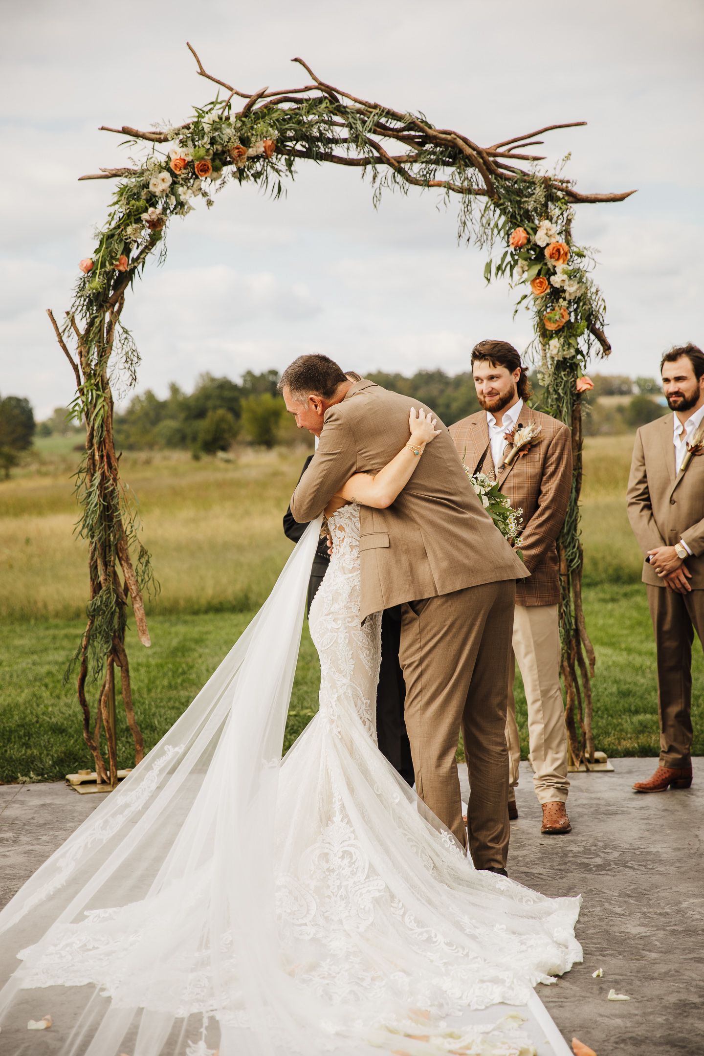 Wedding ceremony under rustic floral arch with brown-suited groomsmen.