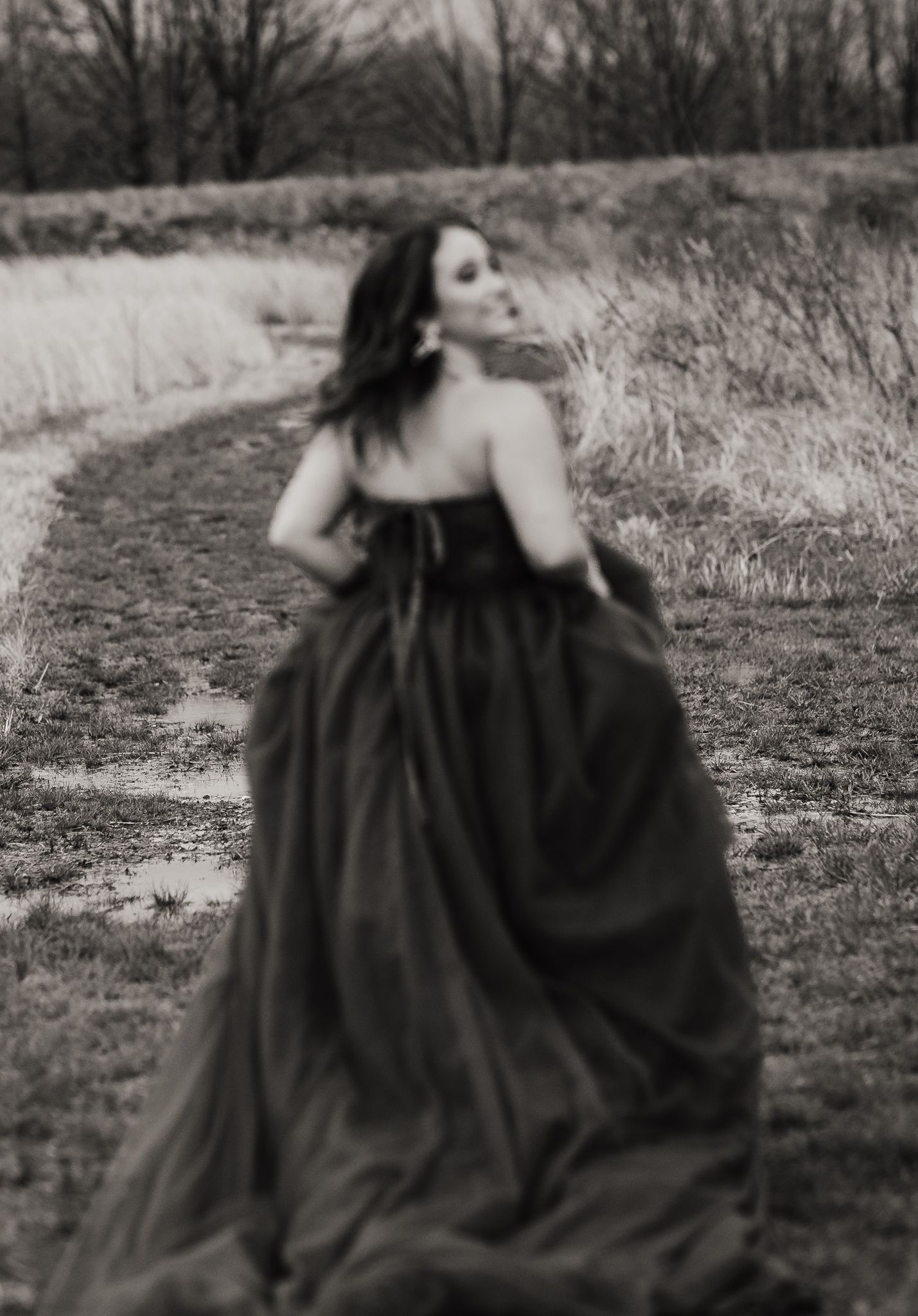 A dramatic black and white portrait of someone twirling in a flowing dark gown on a rural path.