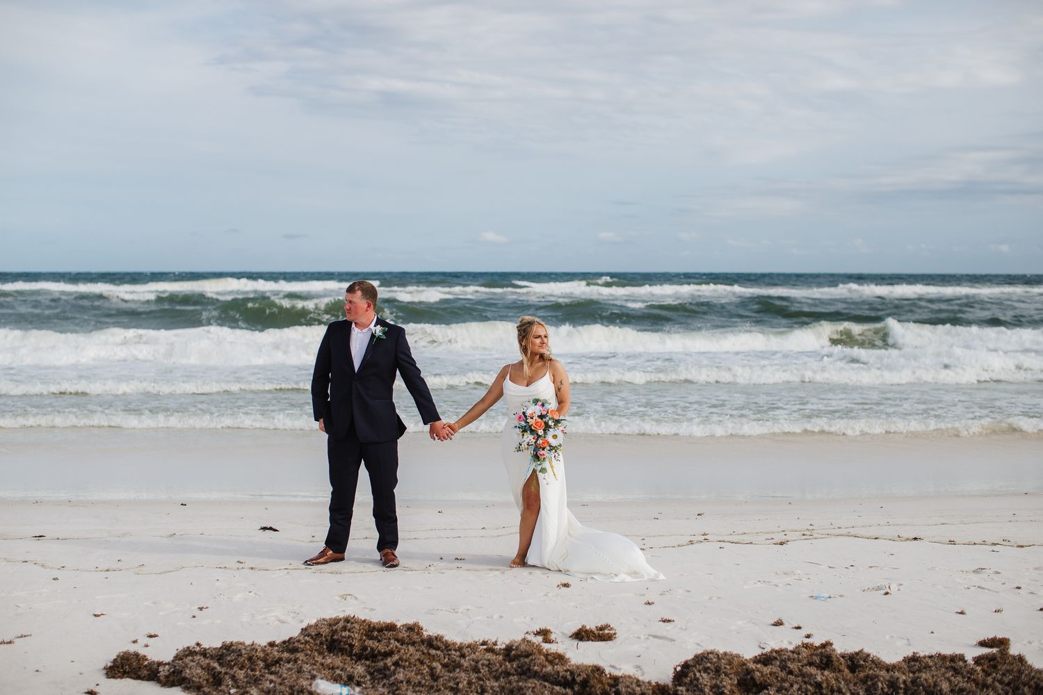 A couple walking hand in hand on a beach with crashing waves in the background.