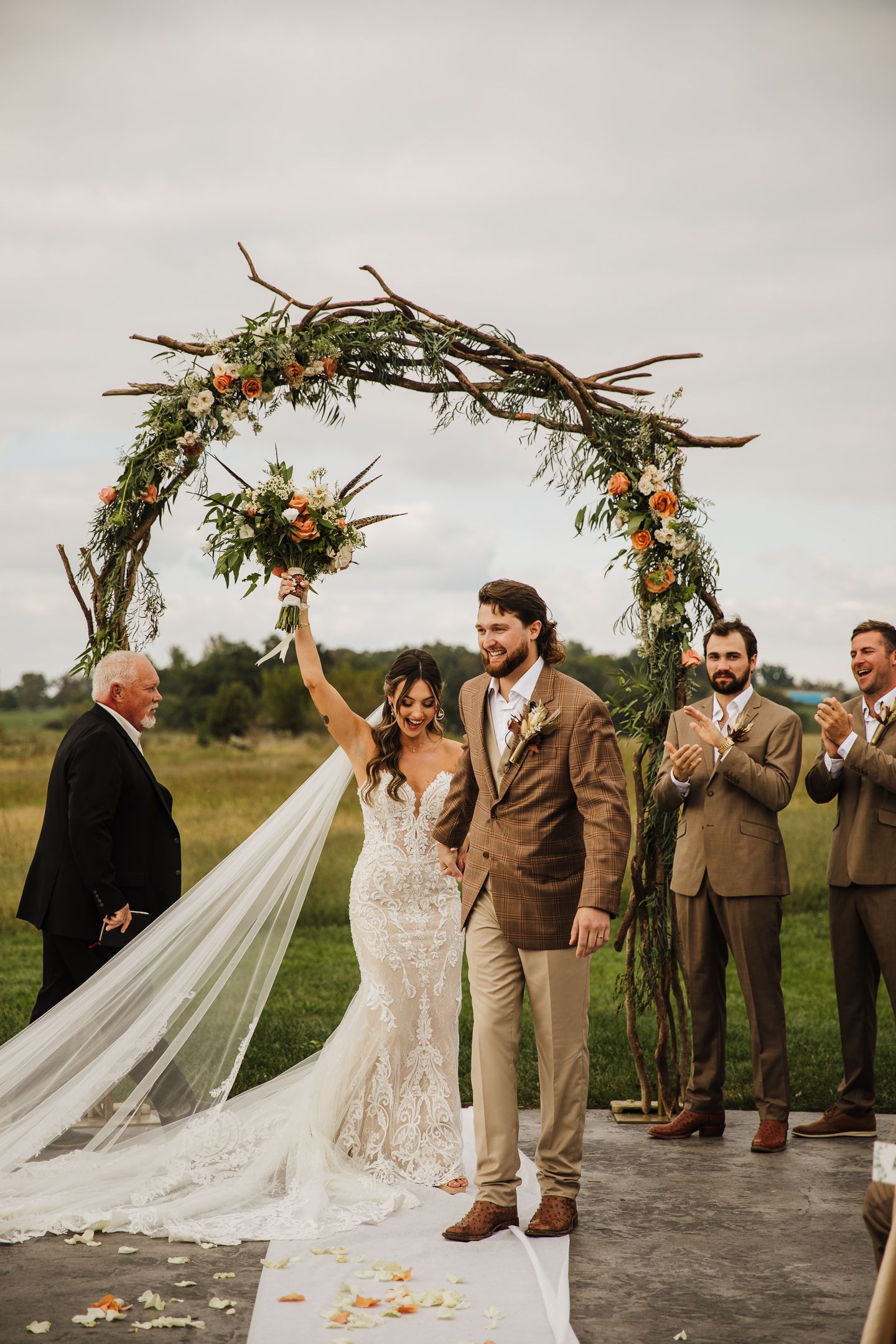 Wedding ceremony under a rustic wooden arch decorated with autumn flowers and greenery in an outdoor setting.