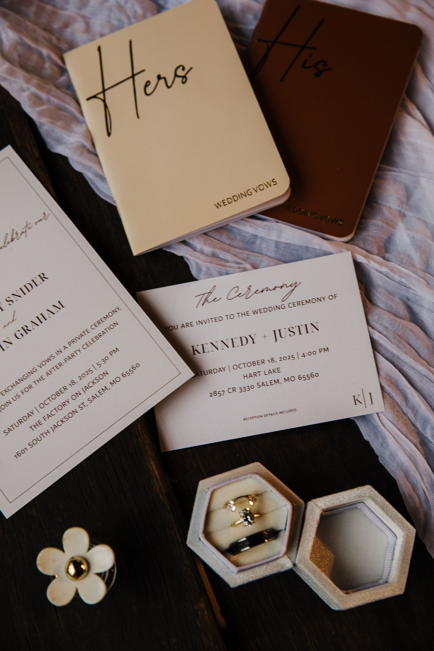 Wedding invitation suite with rings displayed in hexagonal boxes on textured fabric.