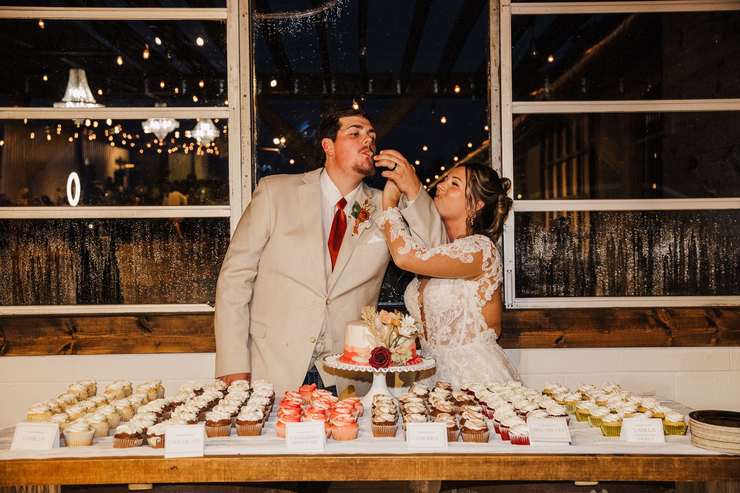 Wedding couple cutting cake together at reception with string lights in background.