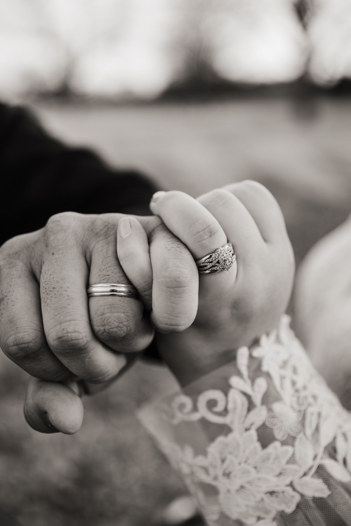 Black and white close-up of two hands with wedding rings touching, with delicate lace detail visible in the background.