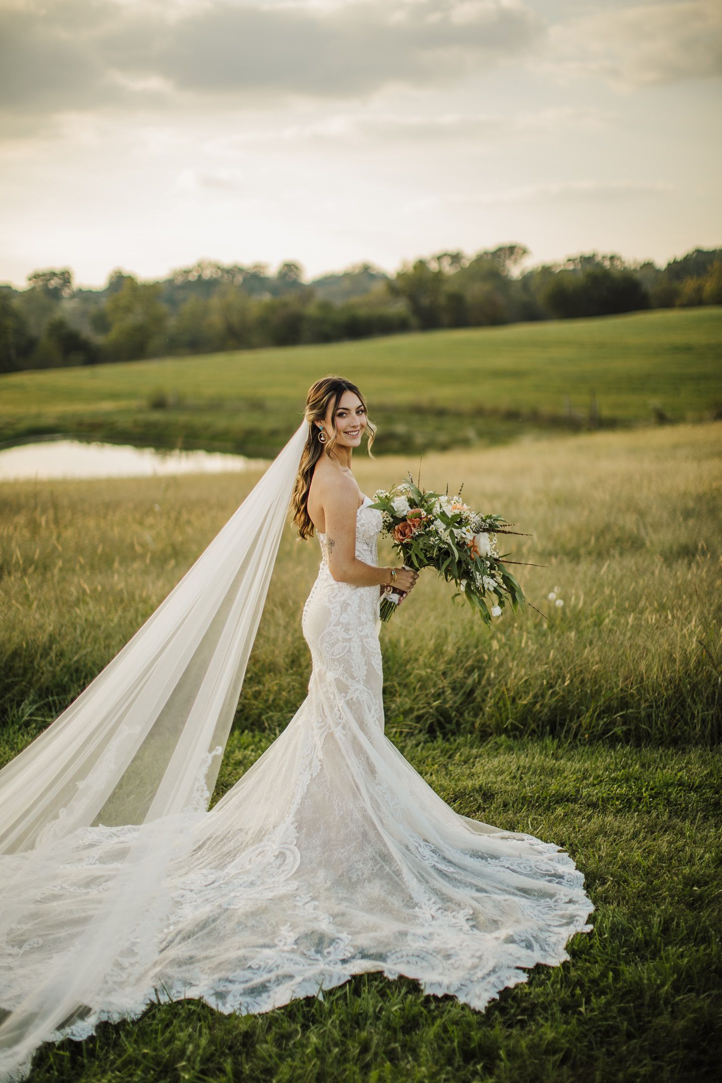 A beautiful lace wedding dress with long train flows across a green field at sunset.