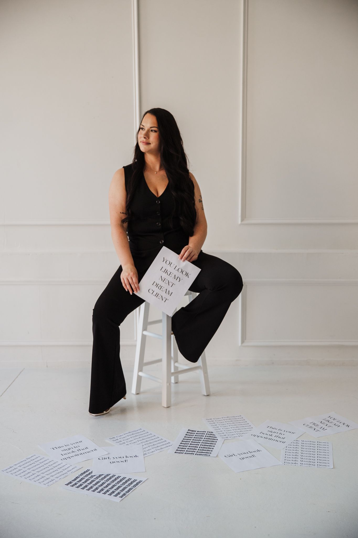 Woman in black outfit arranging papers on white floor in minimalist studio setting.