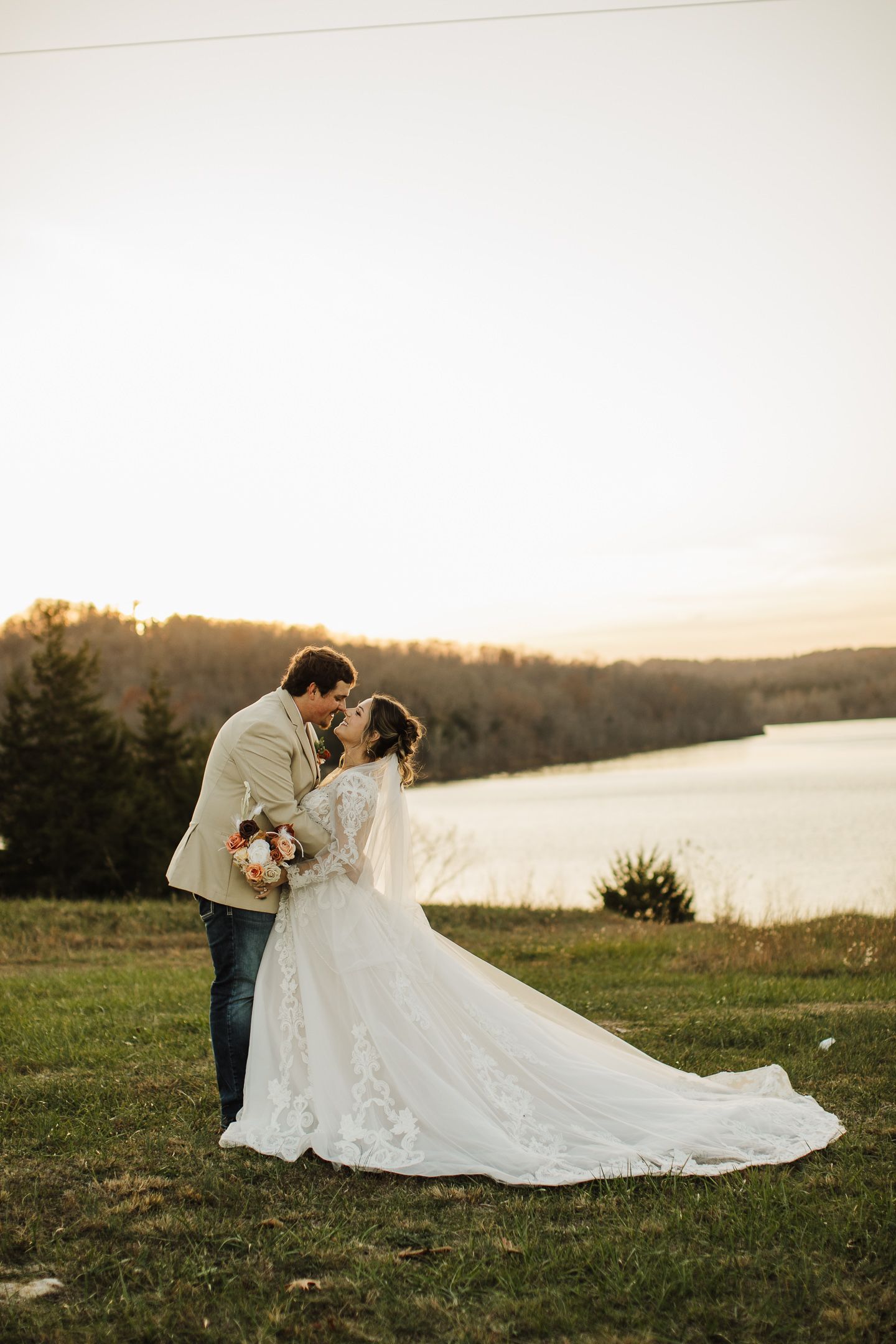 A couple poses for romantic wedding photos at sunset by a lake, with the bride's dress train flowing in the grass.