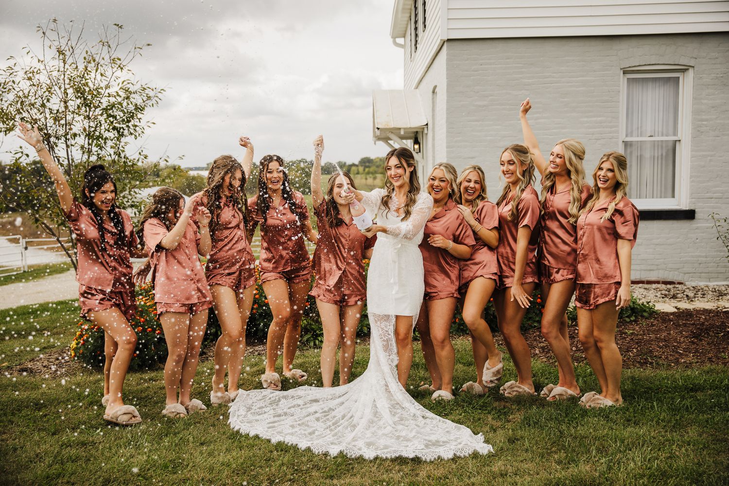 Wedding party in coral dresses poses playfully in a line on grassy lawn outside white building.
