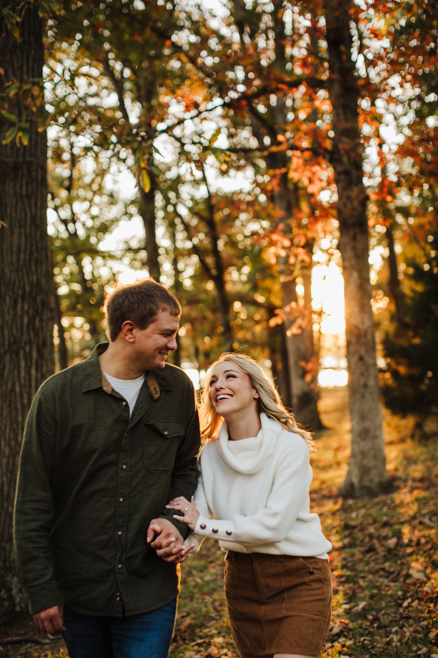 A couple laughs together during an autumn photoshoot with golden sunlight filtering through trees.