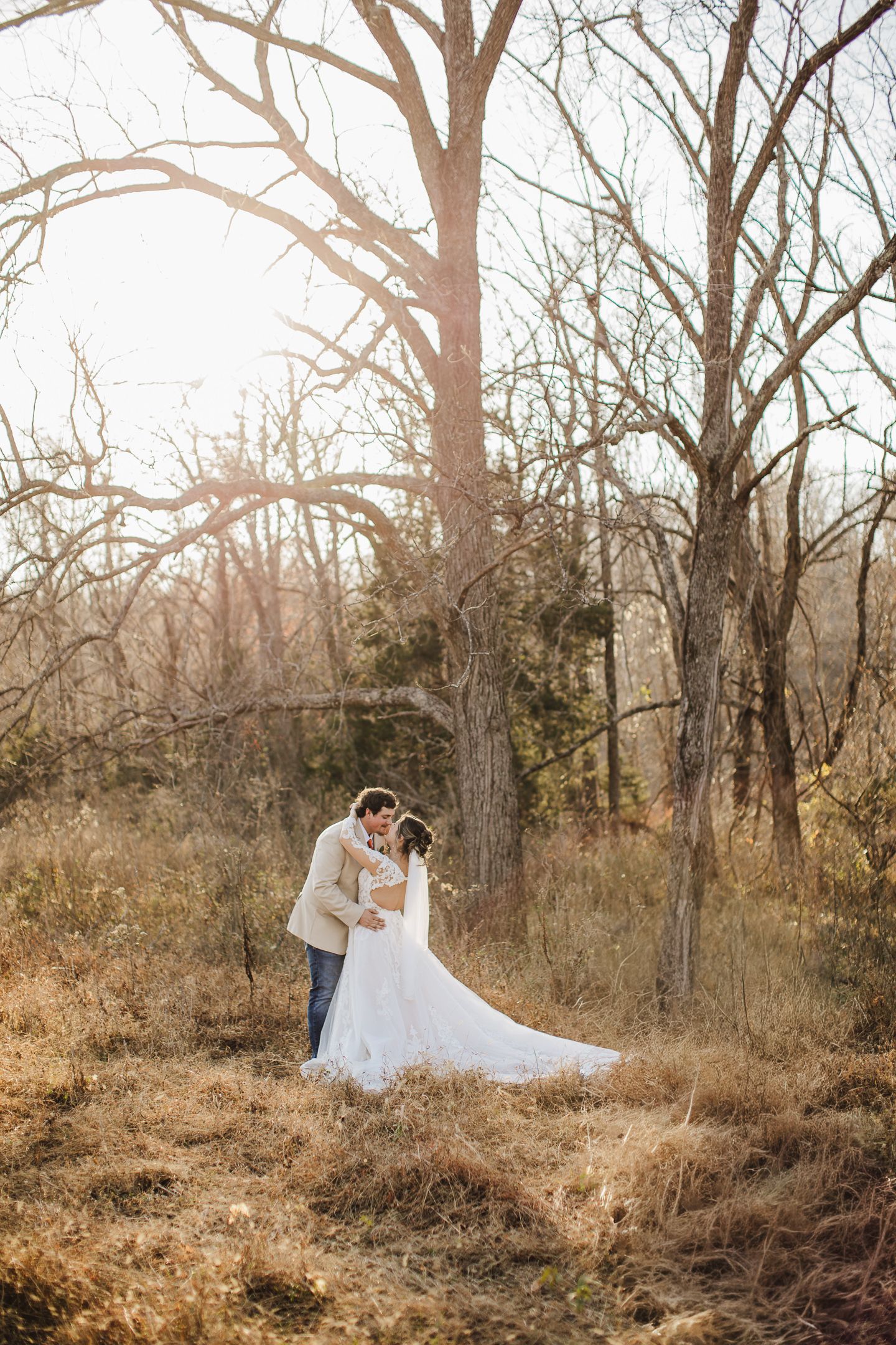 A bride in a flowing white dress poses gracefully among bare winter trees in a rustic outdoor setting.