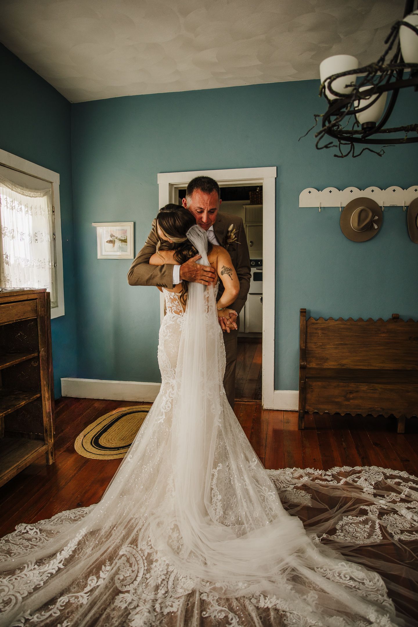 A stunning lace wedding dress with a dramatic train is showcased in a room with teal walls and hardwood floors.