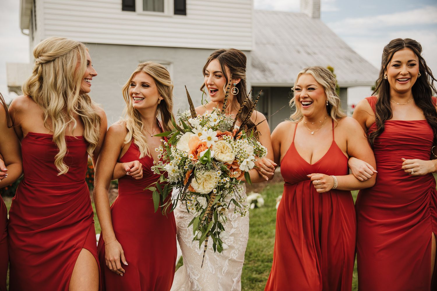 Bridesmaids in burgundy dresses laugh together outdoors holding floral bouquets.