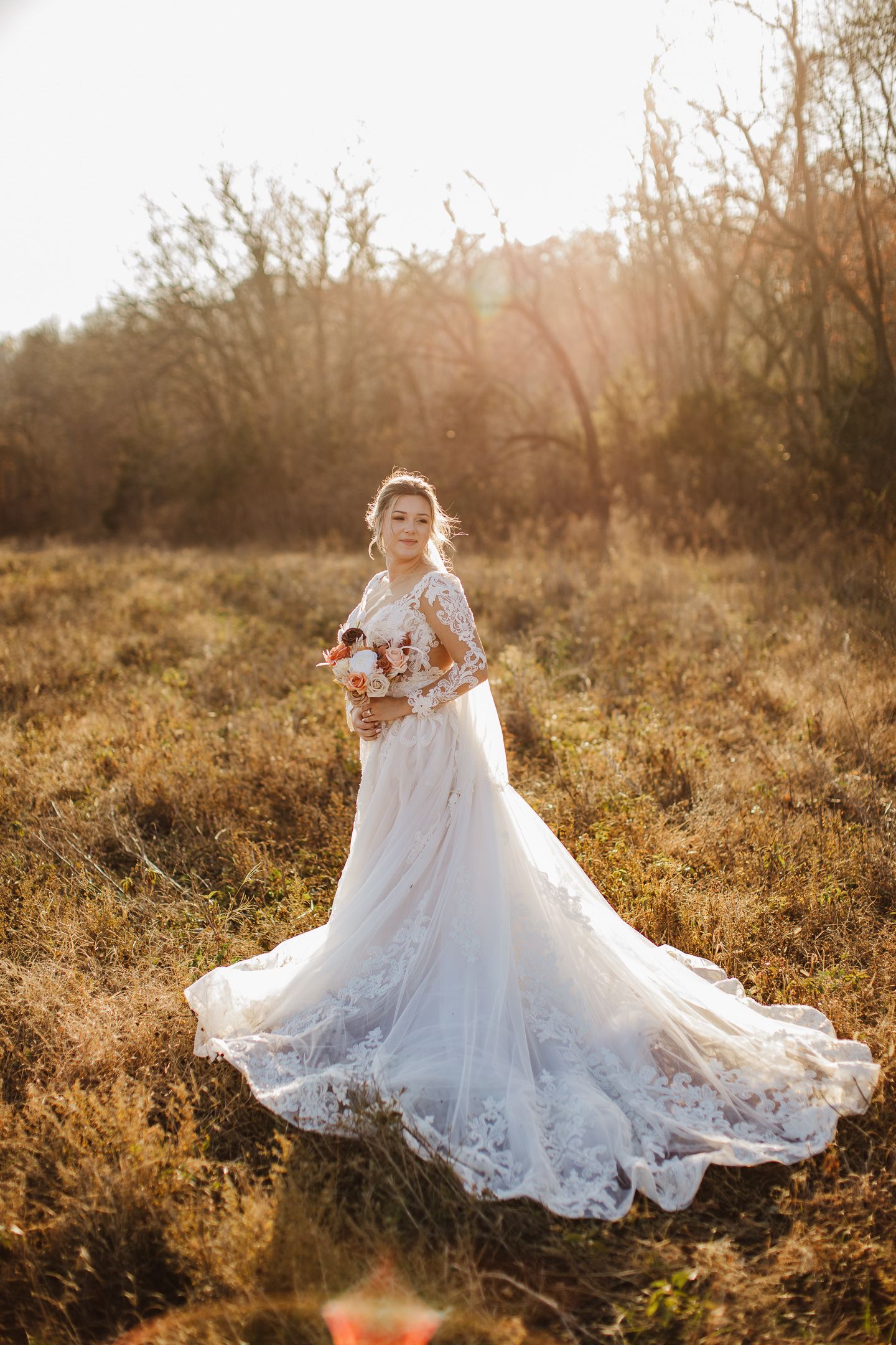 Elegant wedding dress with intricate lace details and flowing train photographed in golden sunlit field at sunset.