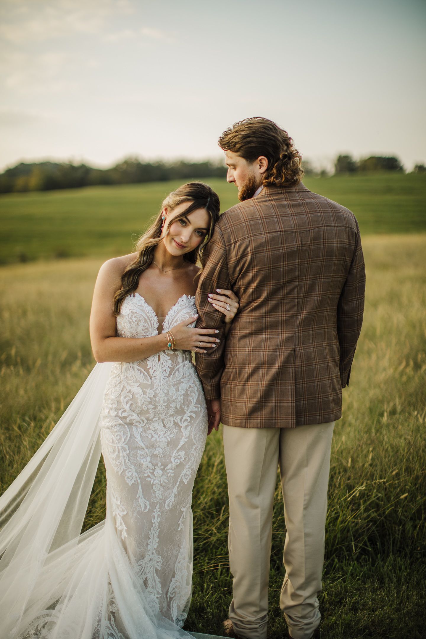 A tender wedding moment captured in a rustic outdoor setting with flowing white dress and brown plaid suit.