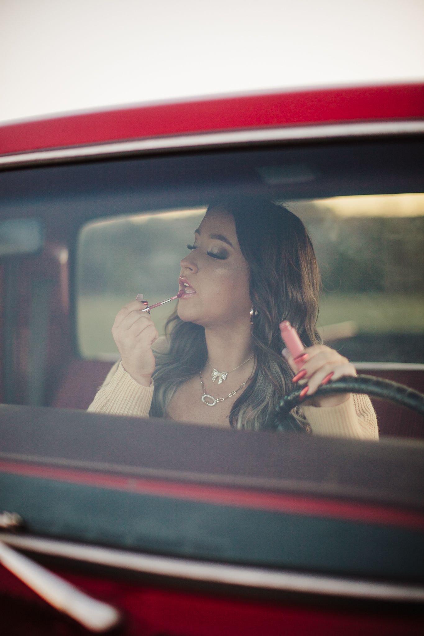 Woman applying lipstick while seated in classic red car with vintage styling.