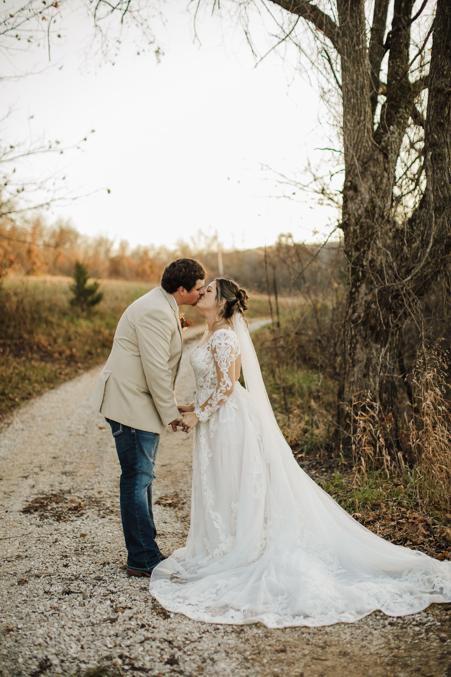 A couple in wedding attire share an intimate moment on a gravel path surrounded by autumn trees.