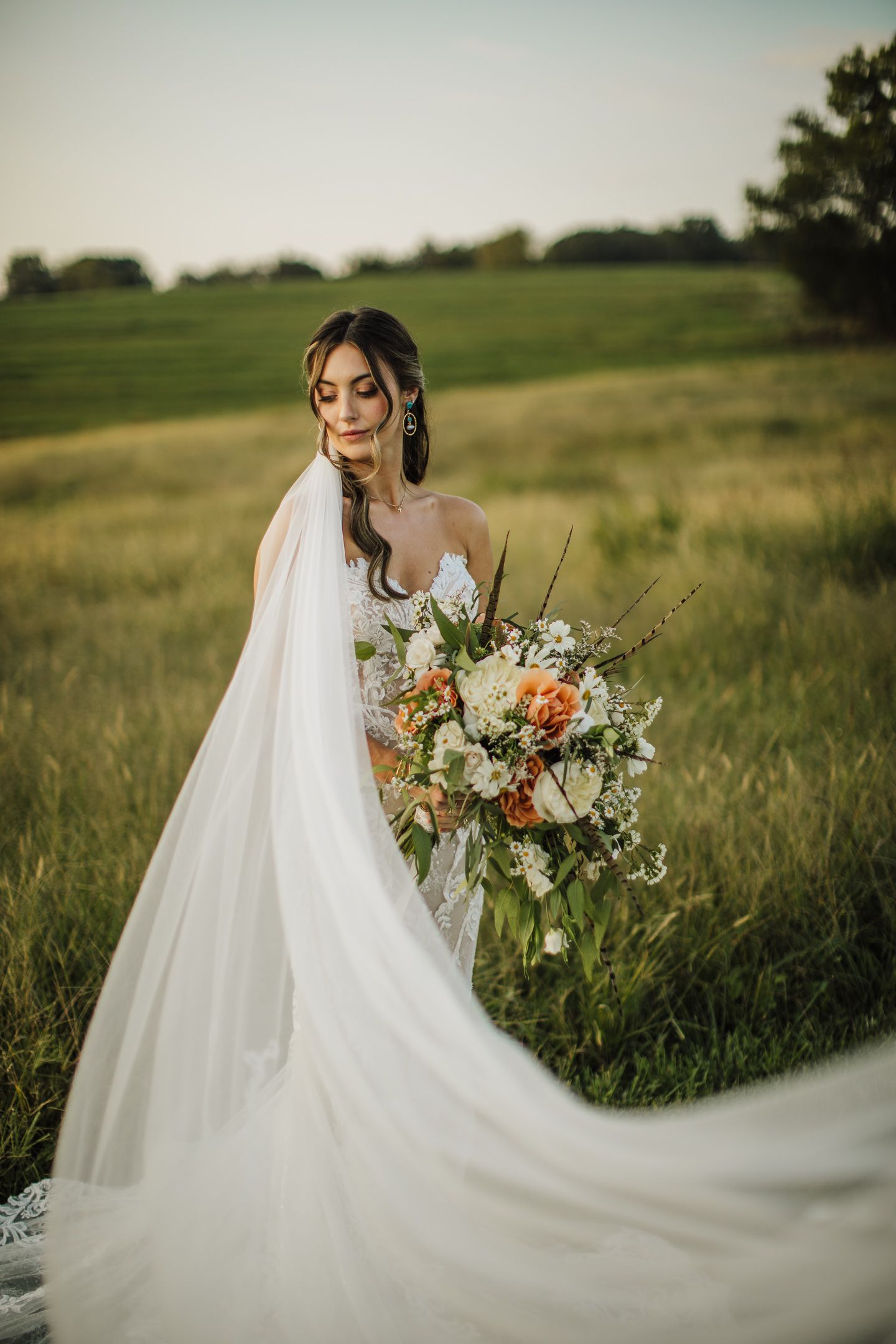 Bride's long veil flows dramatically in wind against green field backdrop.