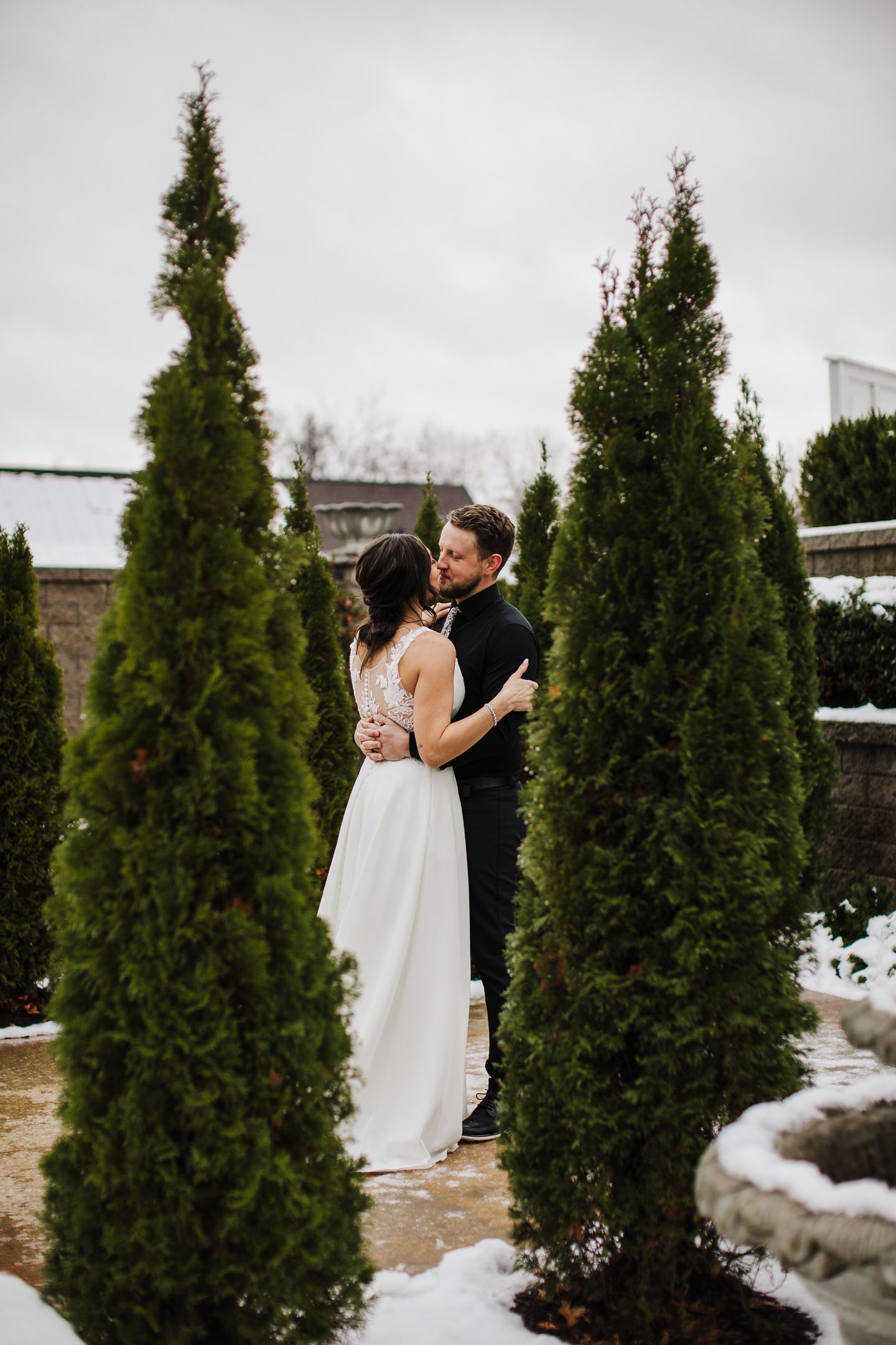 A couple shares a romantic moment among snow-covered evergreen trees in a winter garden setting.