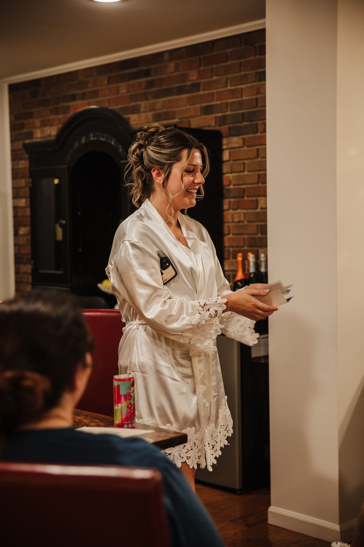 A person in a white lace-trimmed satin robe stands in a room with exposed brick walls and leather seating.