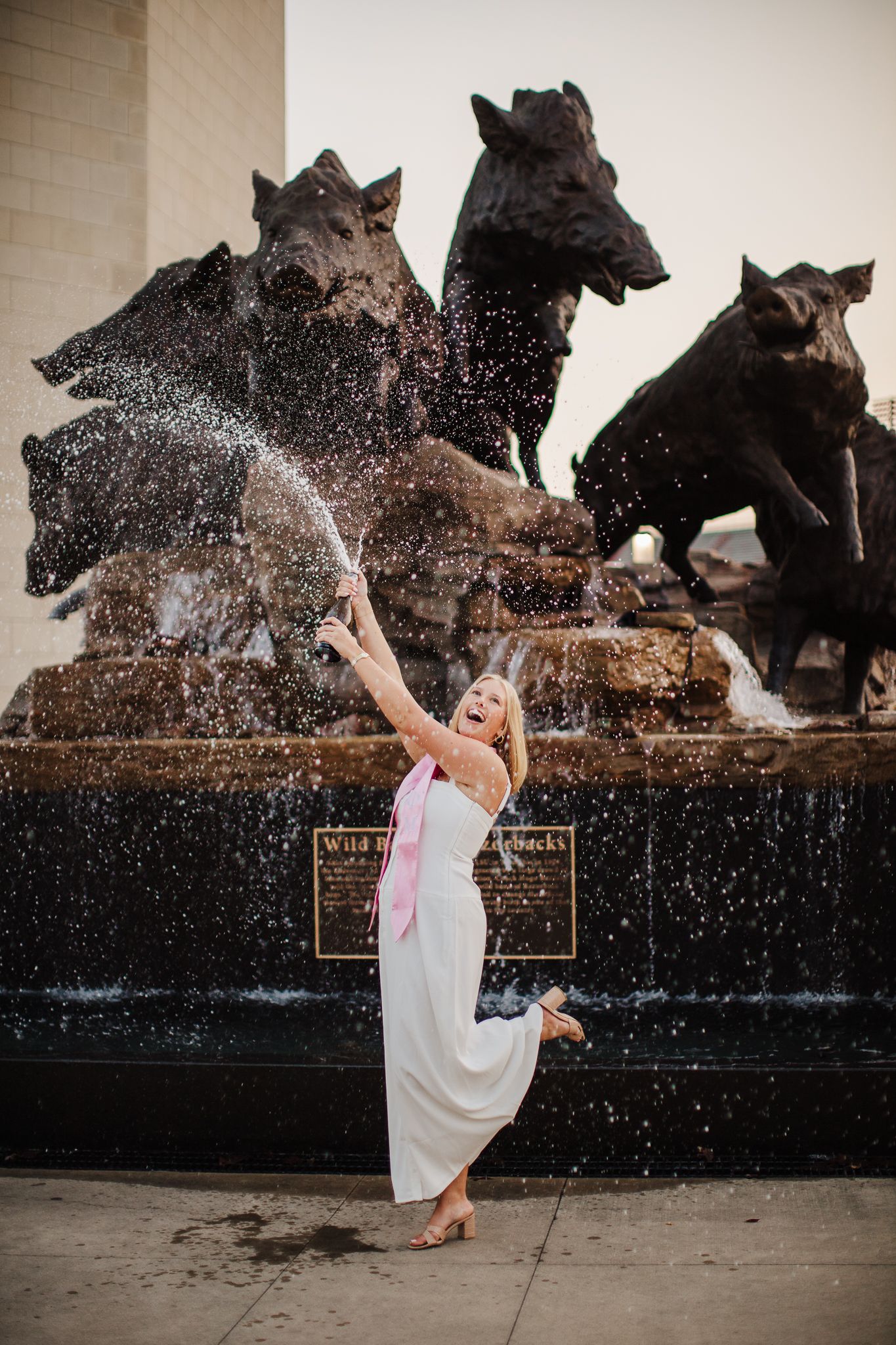 A person in white dress poses playfully in front of a wolf fountain sculpture.