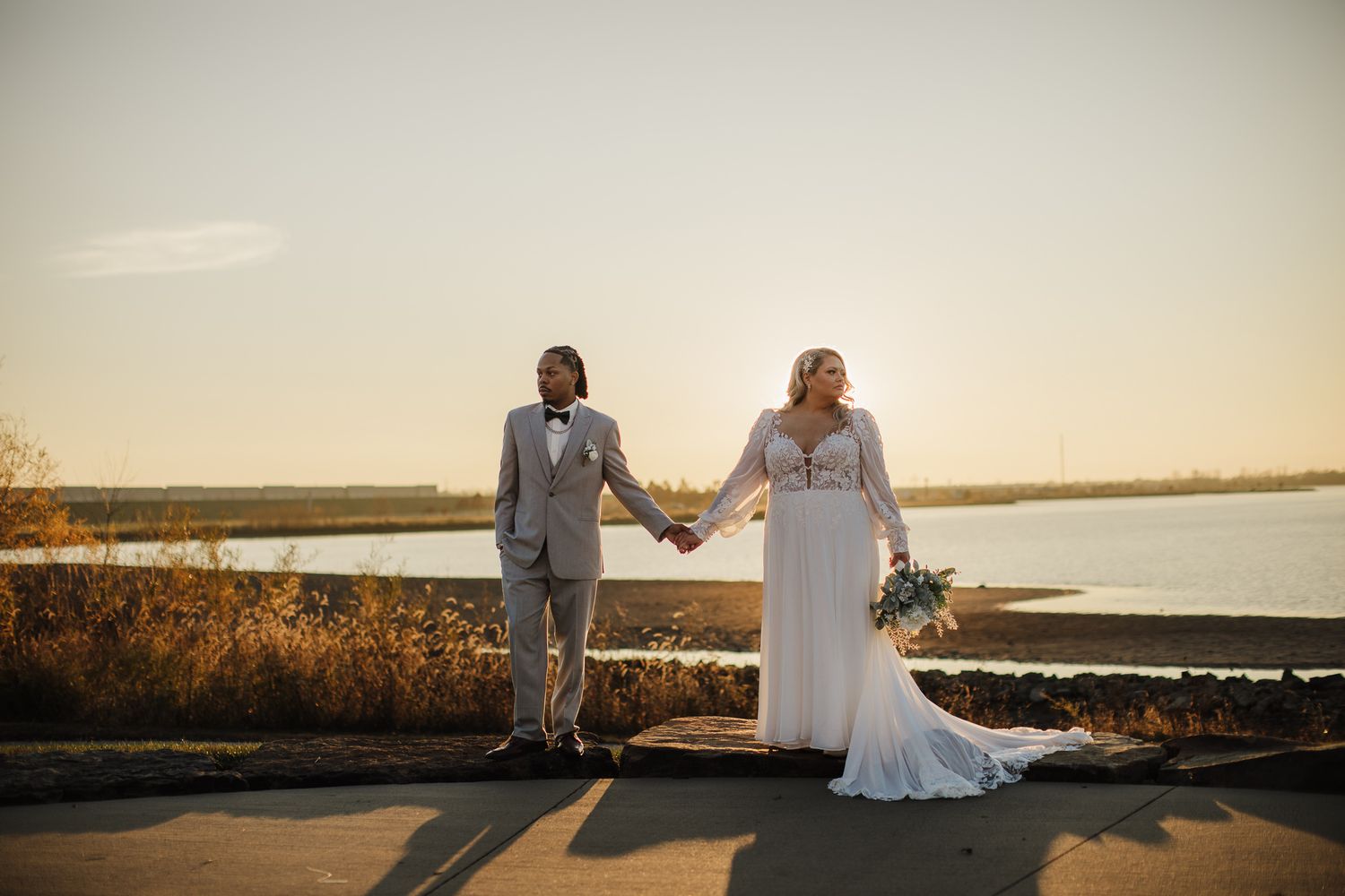 Newlywed couple sits on a wooden dock at sunset overlooking a serene lake with golden light in the background.