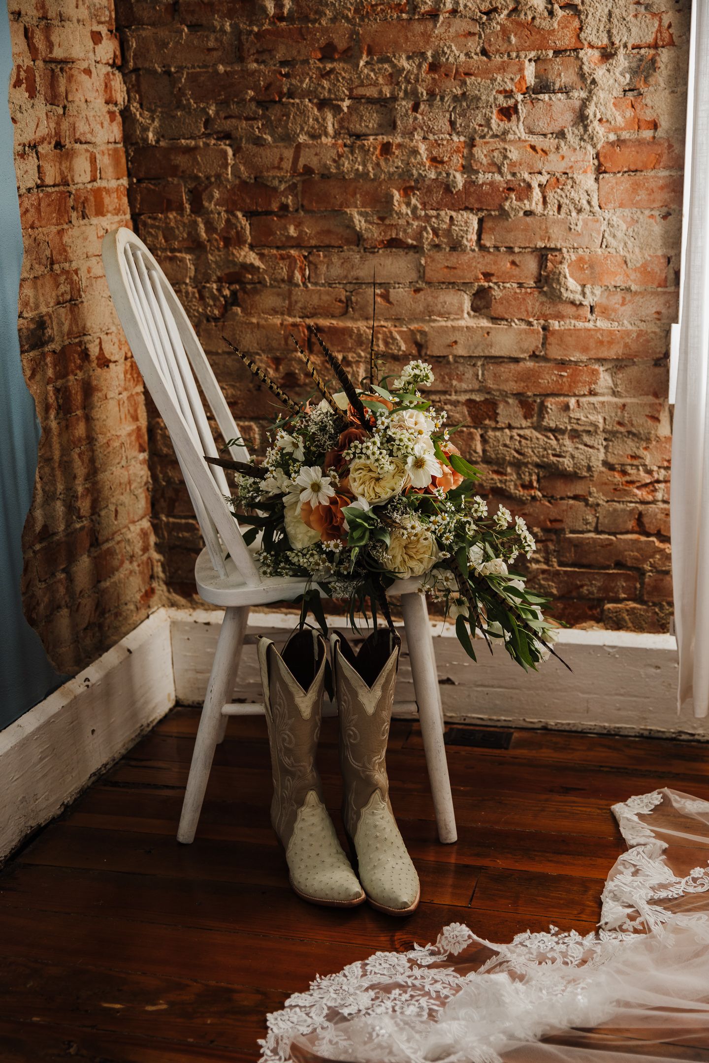 White bridal boots and floral bouquet arranged with lace details against exposed brick wall and wooden floor.
