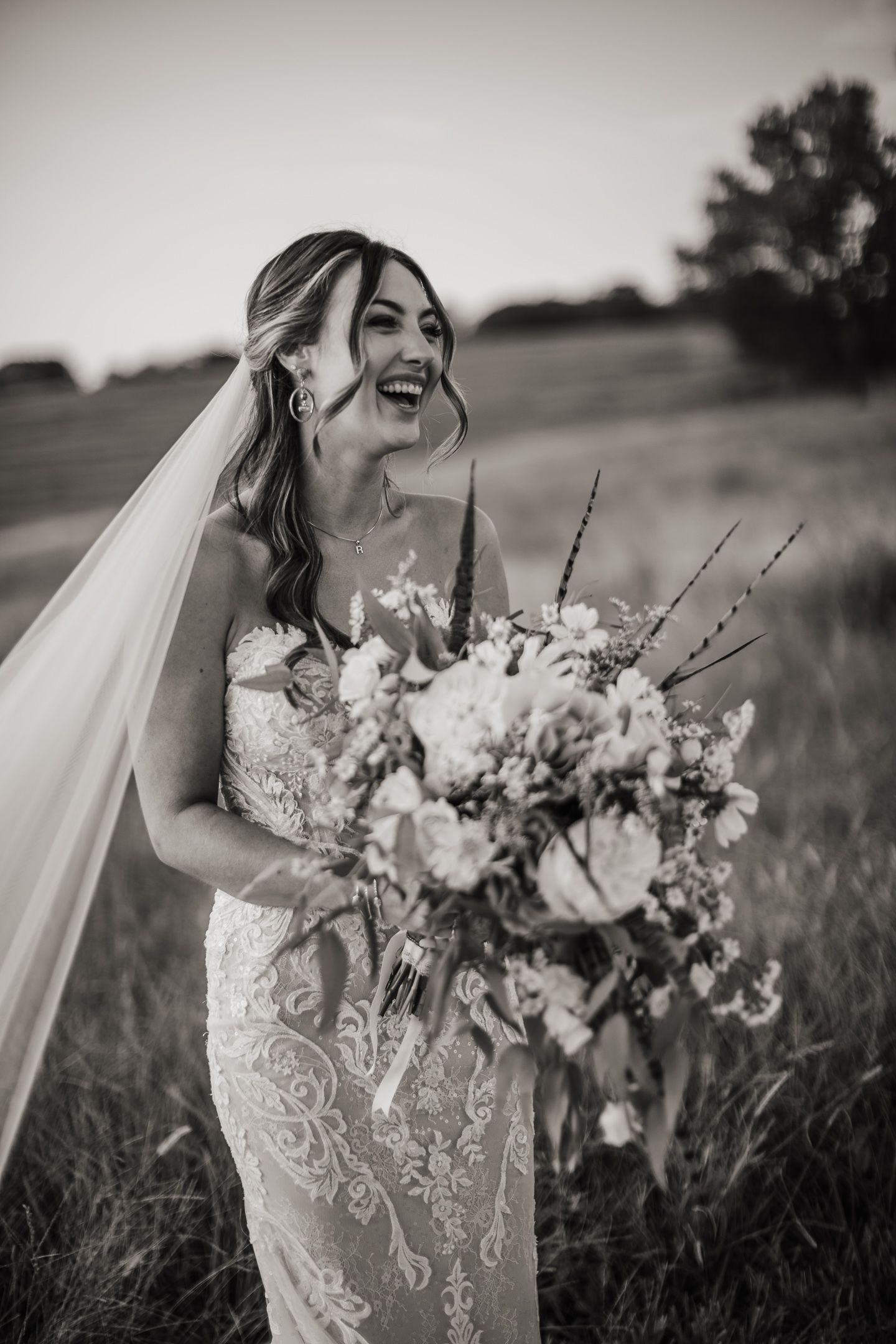 A bride laughs while holding a stunning white and purple floral bouquet in a field.