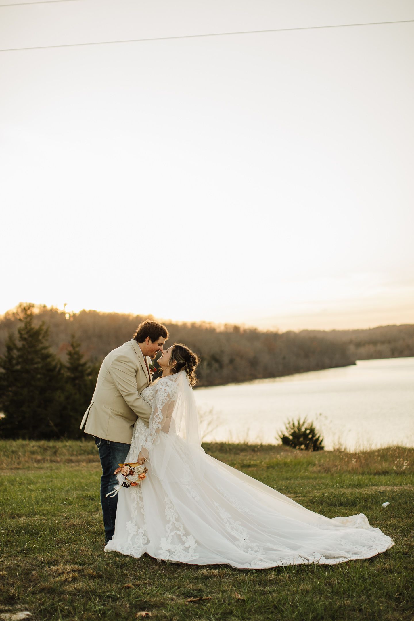 A newlywed couple embraces during golden hour by a lake with the bride's lace dress trailing behind them.