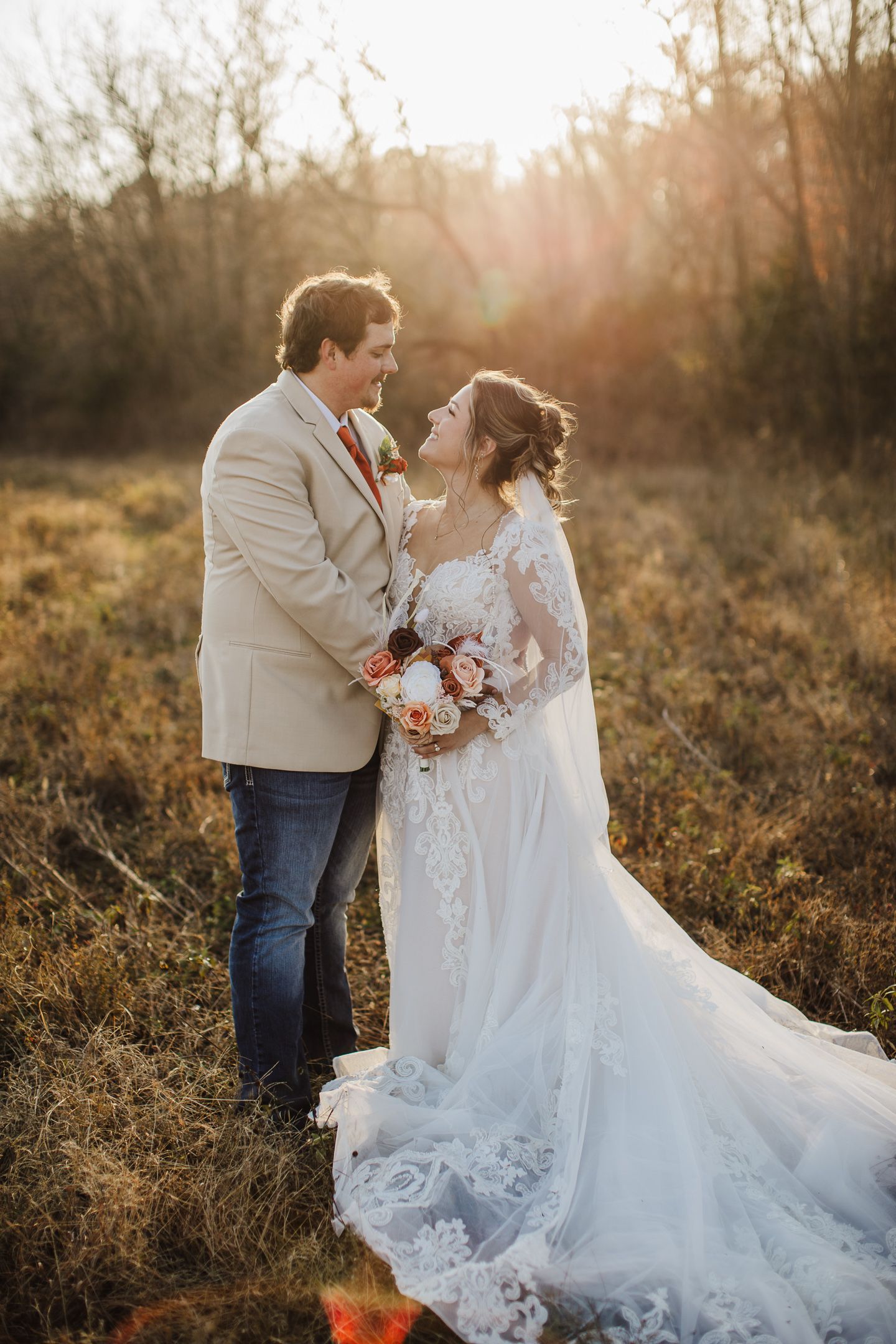 Couple in wedding attire embrace at sunset in a golden field with bride wearing lace gown and groom in beige blazer.