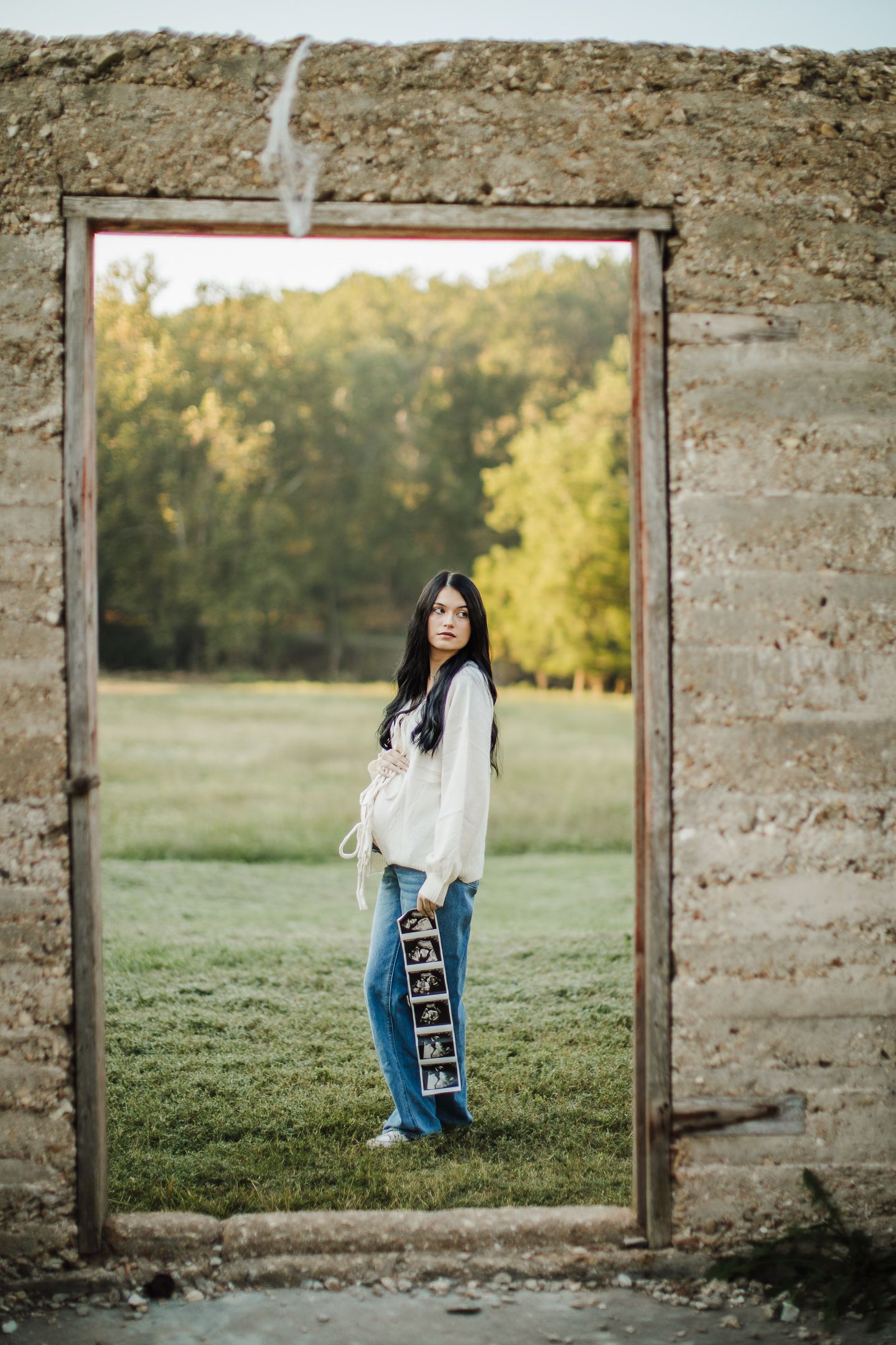A maternity portrait framed within a rustic stone doorway overlooking green fields.