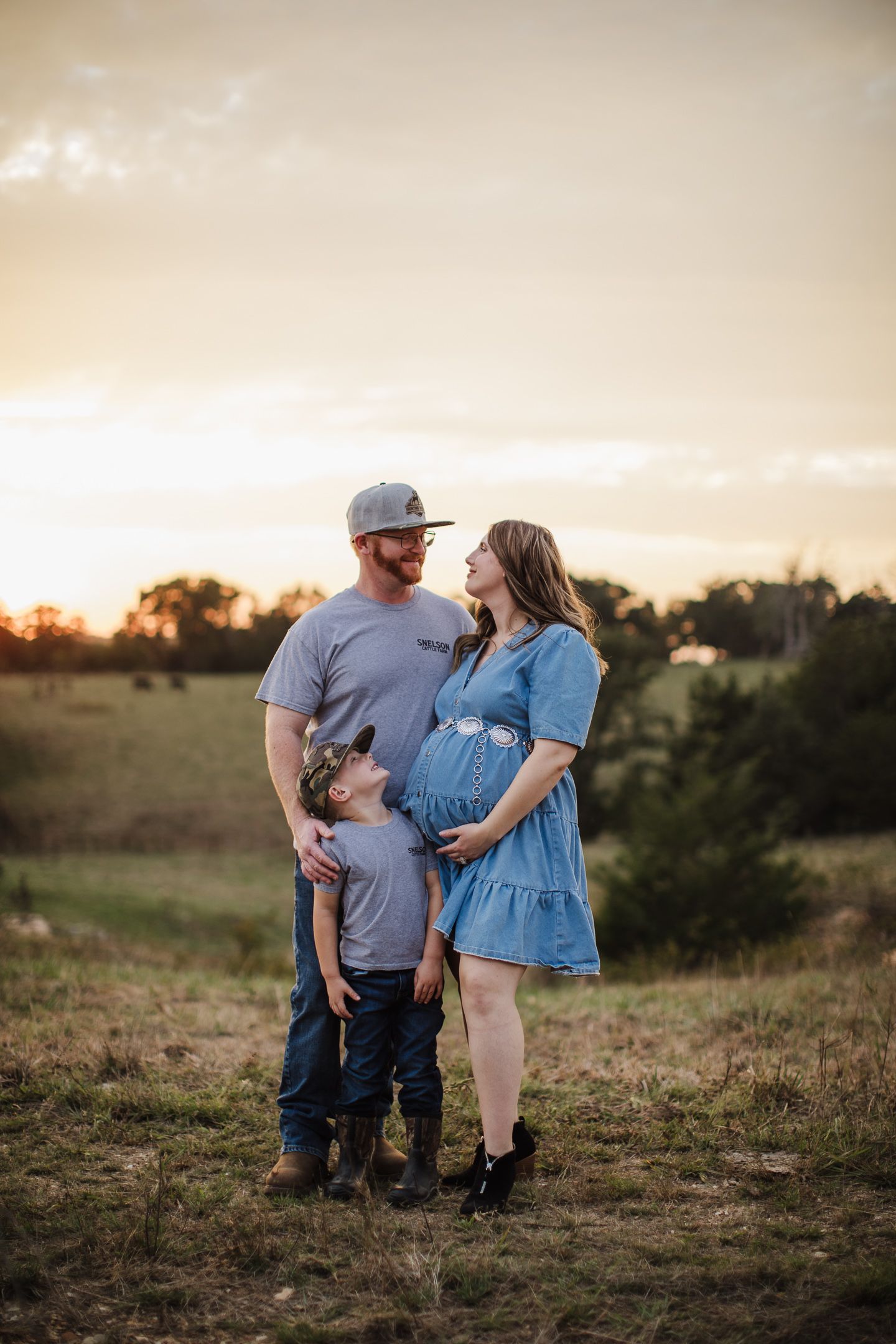 A sunset maternity photo of a family in casual clothing on a grassy hill.