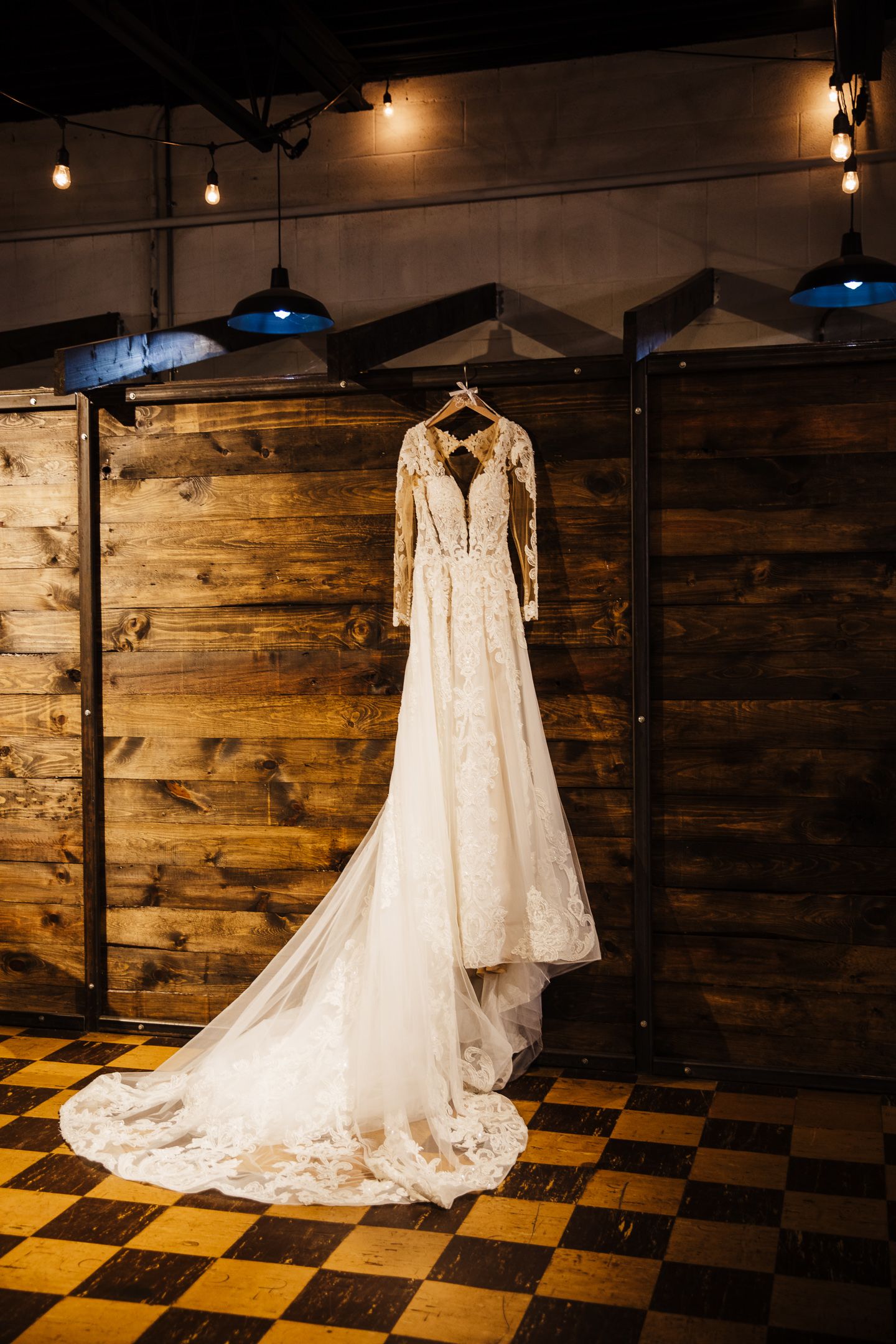 Wedding dress with intricate lace details displayed on wooden wall with checkerboard floor and string lights.