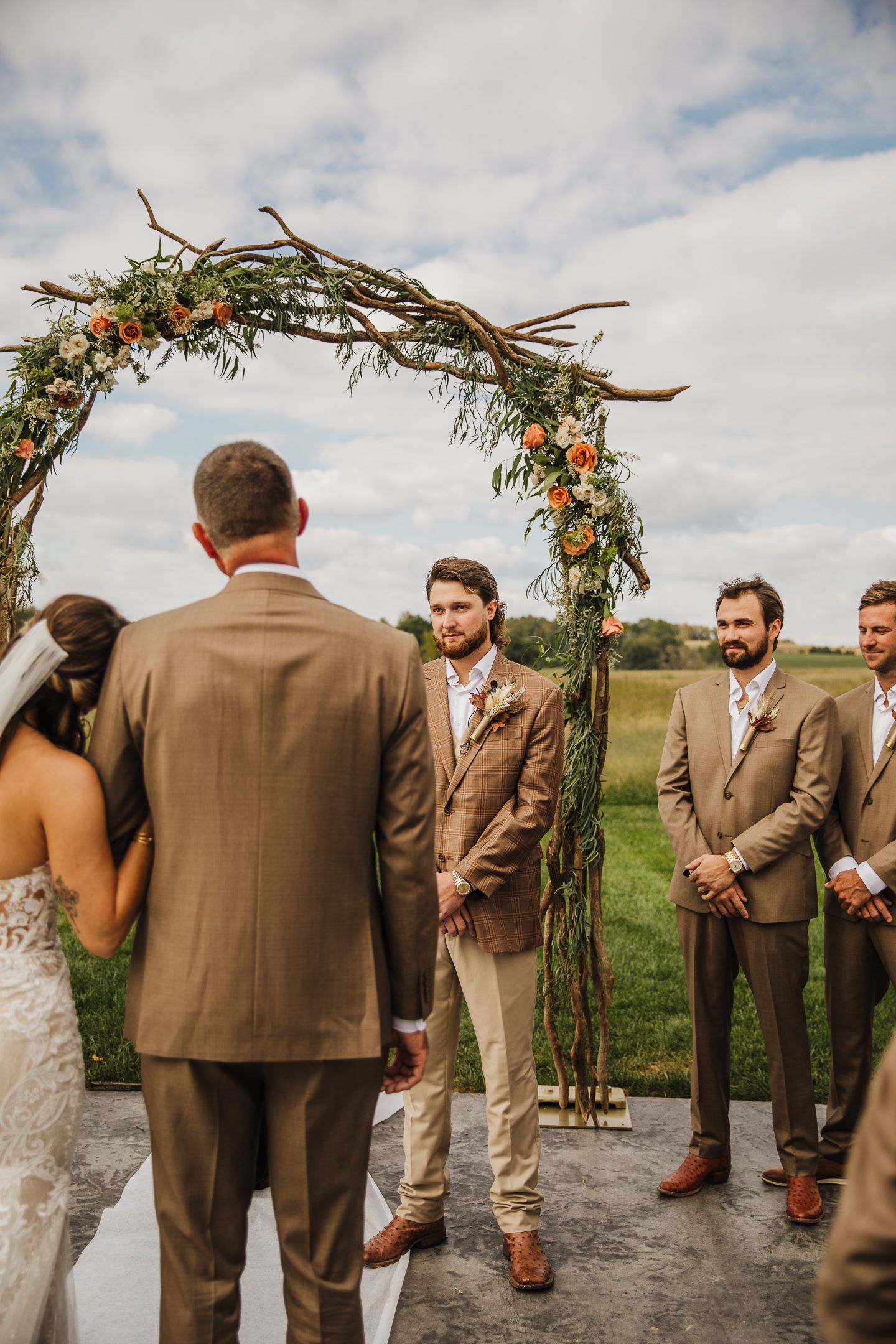 Wedding party stands in front of a floral arch during an outdoor ceremony with brown and tan suits.