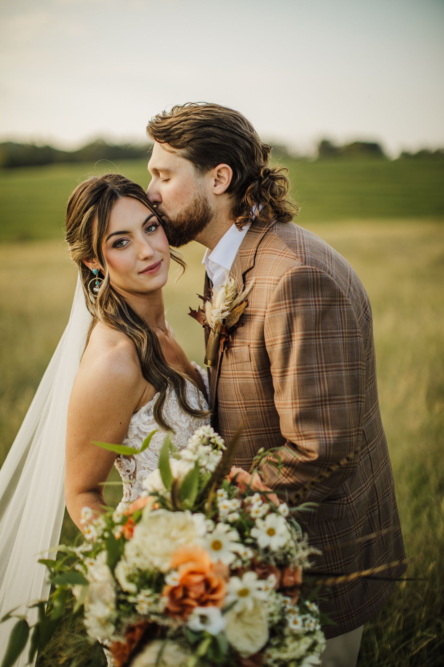 A romantic wedding portrait of a couple in a golden field at sunset with a beautiful floral bouquet.