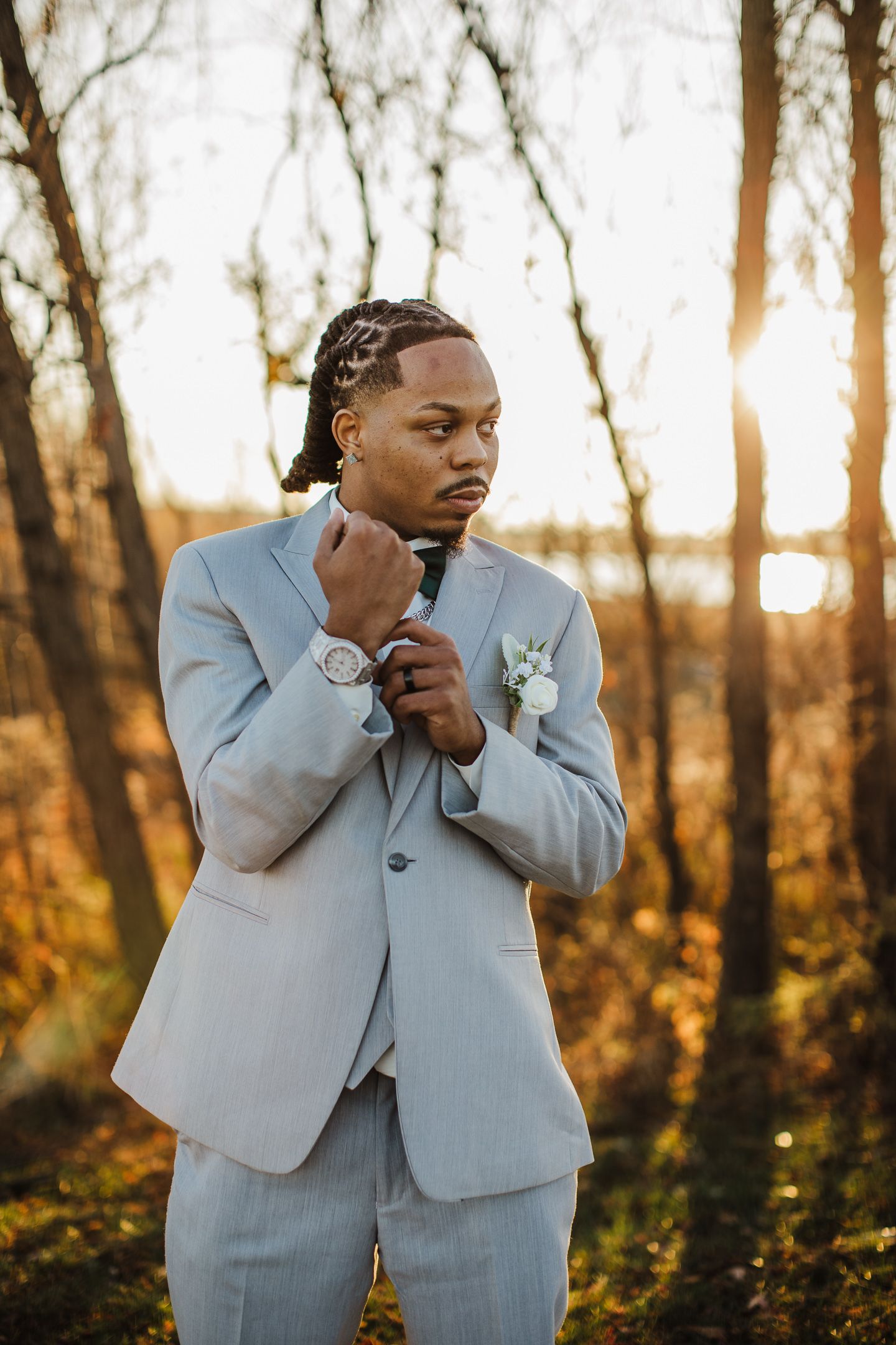 A groom in a light blue suit adjusts his tie in golden sunset light.