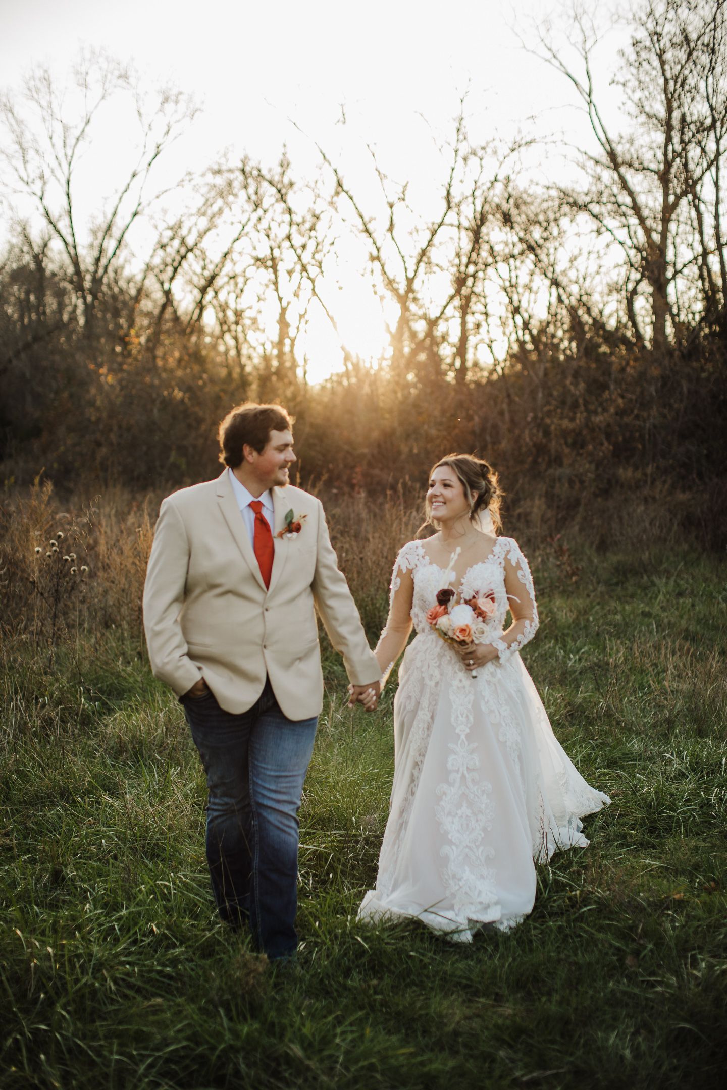 A couple shares romantic moments in a field at sunset, with the bride wearing a lace gown and groom in a beige suit.