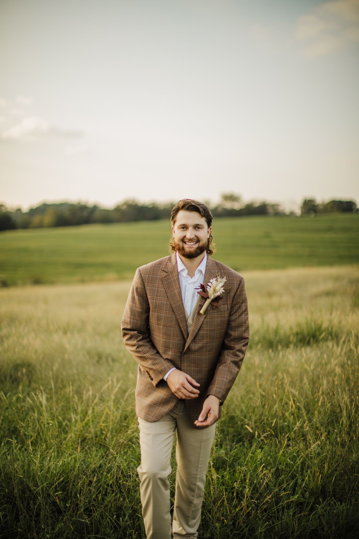 Groom in brown tweed jacket poses in golden field during sunset.
