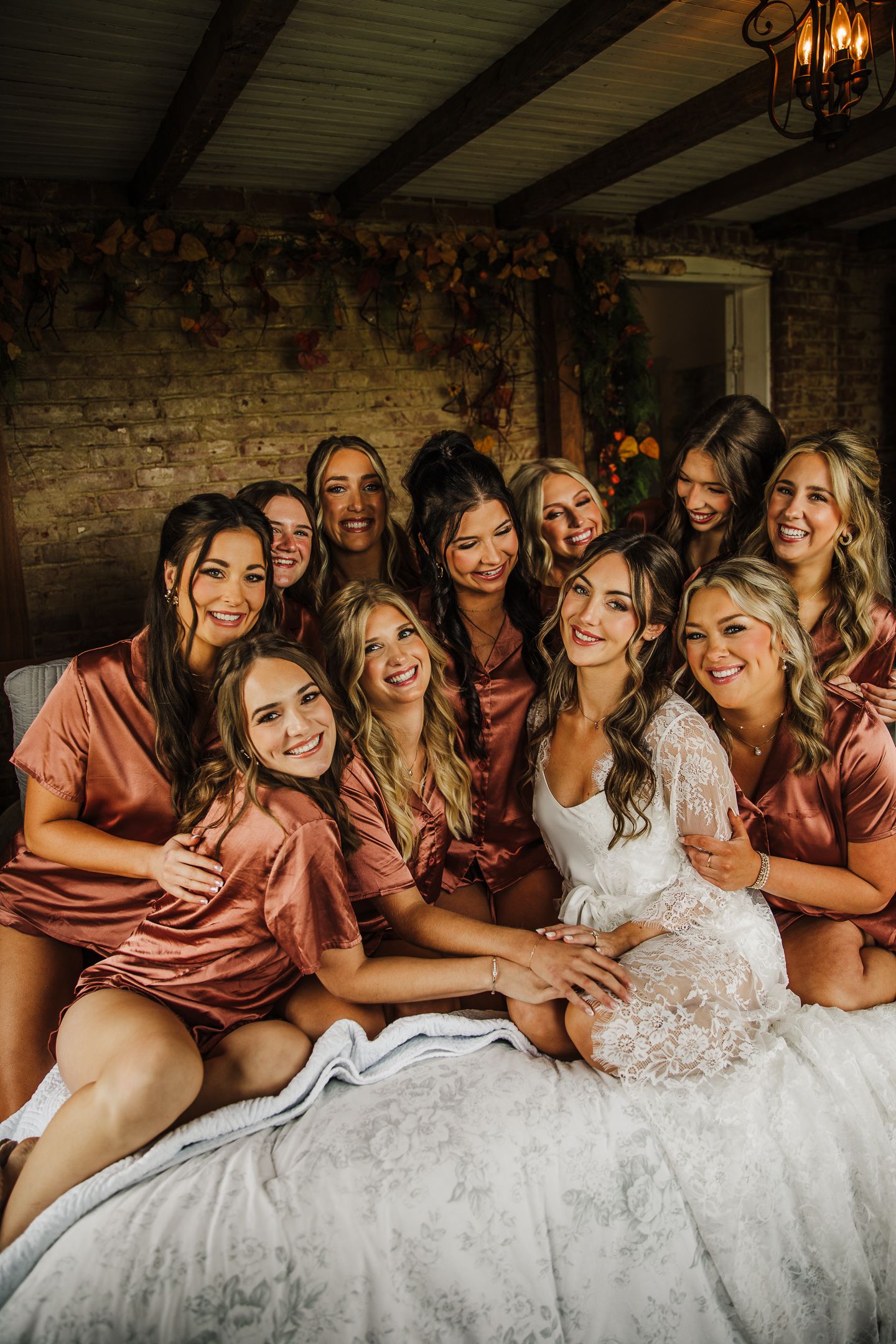 A bridal party in silk robes lounges together on a bed sharing laughter and smiles in a rustic room.
