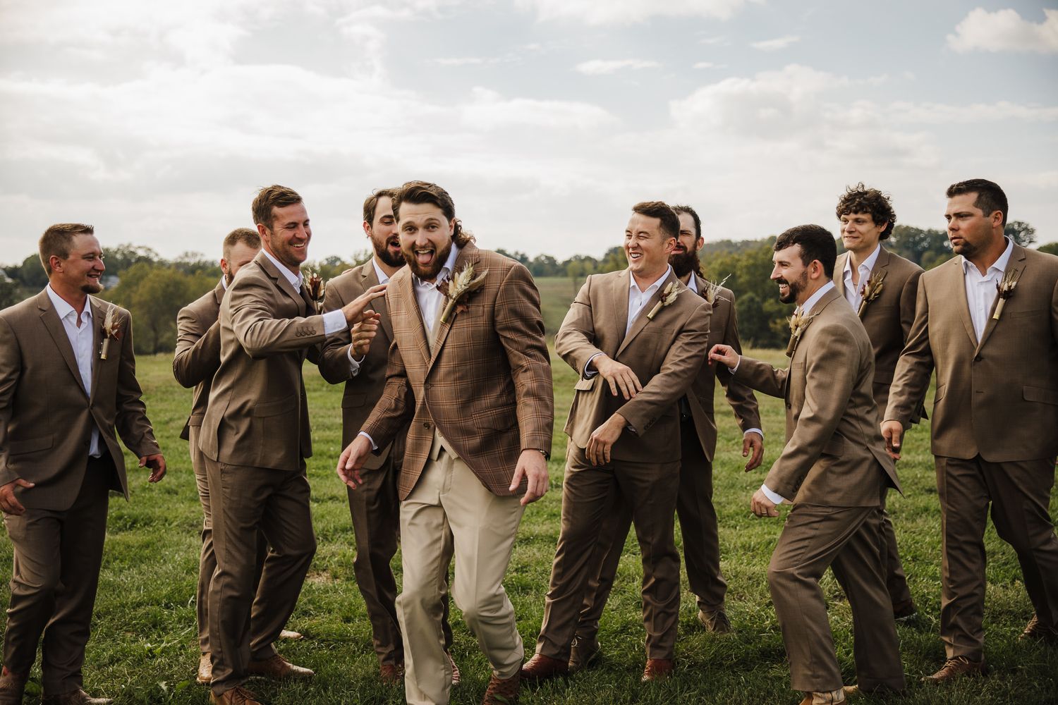 Groomsmen in tan suits pose playfully in outdoor field setting.