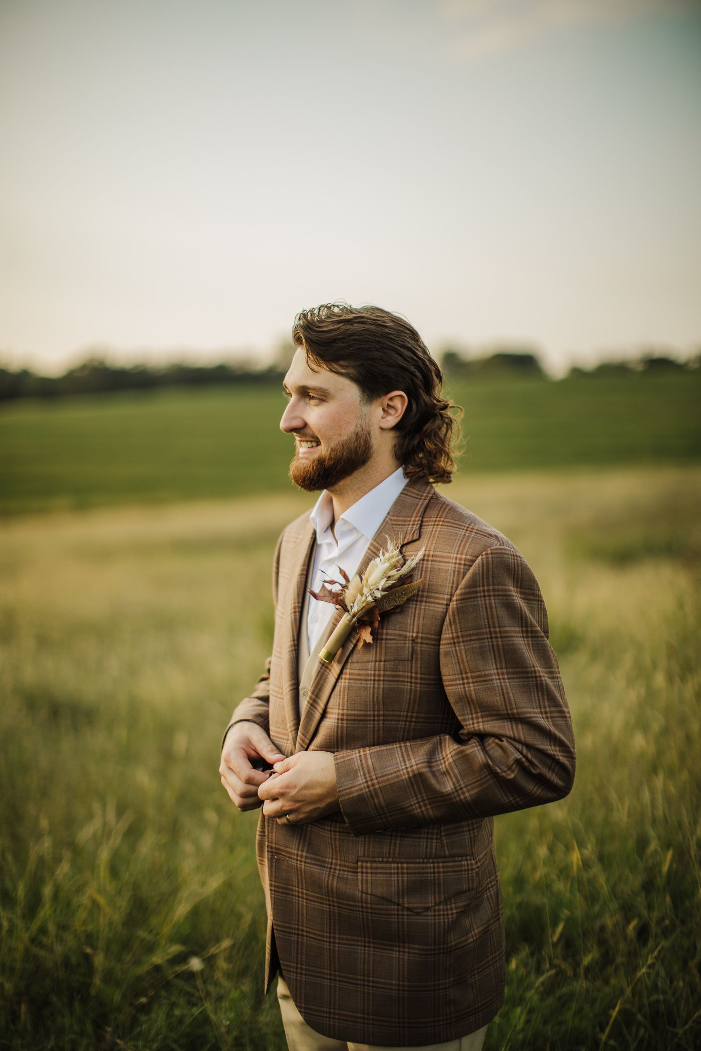 A person in a brown plaid suit adjusts their boutonniere while standing in a sunlit grassy field.